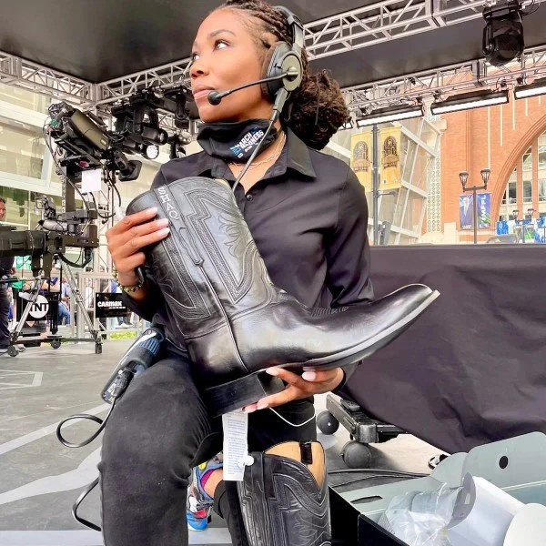 A woman in a headset holds a large black cowboy boot at an outdoor event with stage equipment and buildings in the background.