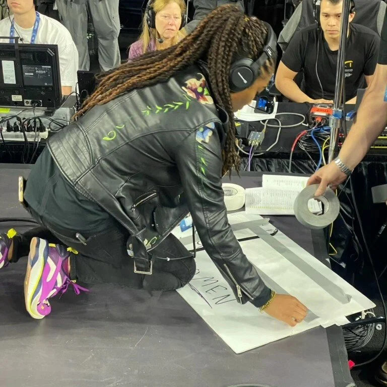 Woman with long braids wearing headphones and a leather jacket, kneeling on a table, working on a large sheet of paper with writing in a crowded media control room with equipment and people.