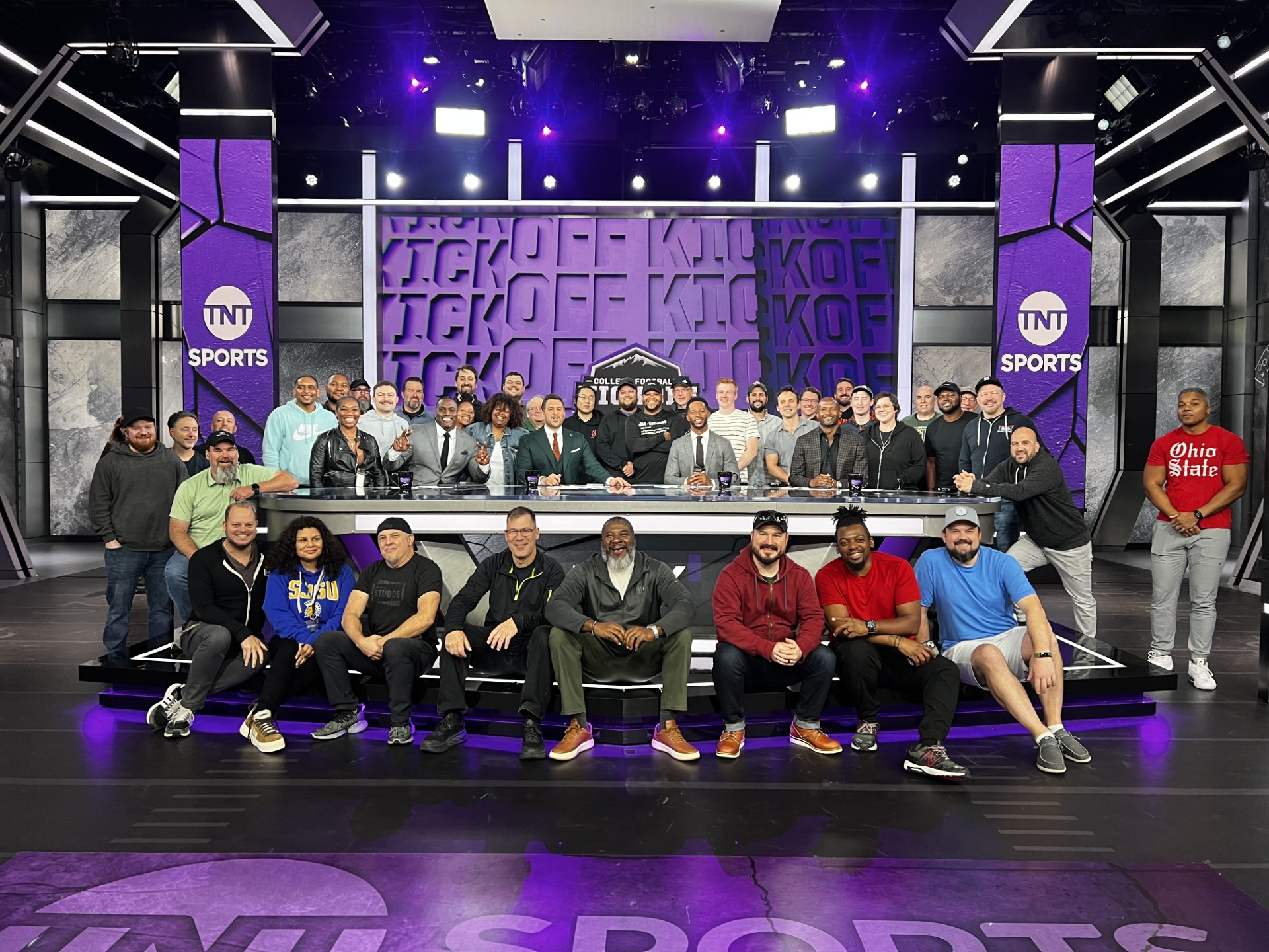 Group photo of diverse sports commentators and analysts on a television set for TNT Sports, with a purple and black stage background, and a large group seated and standing for a team photo.