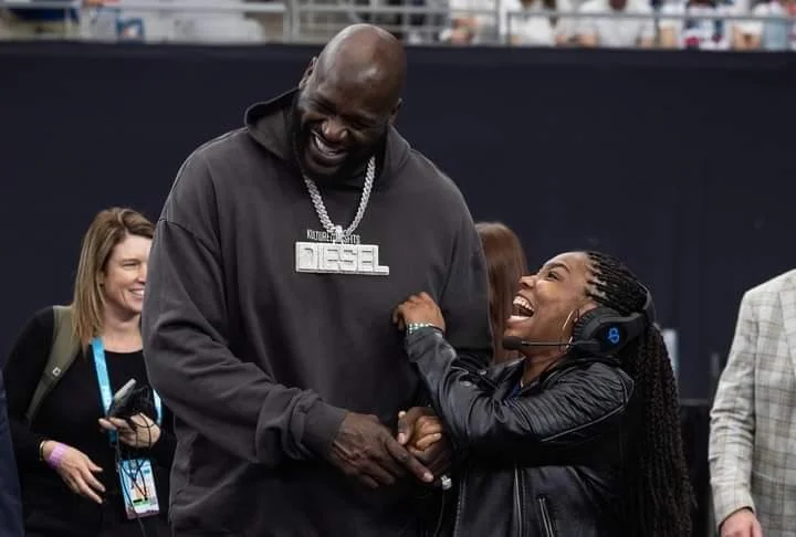 Shaquille O'Neal and Nicole Monet sharing a laugh and smiling while holding hands at an event, with other people in the background.