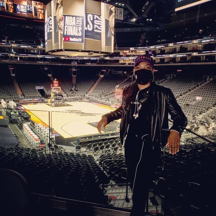 Person wearing a black face mask, dark clothing, and a baseball cap standing inside an empty basketball arena, with the court visible in the background.