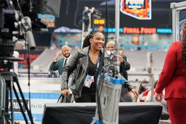 A woman with braided hair smiling and holding a black bag, surrounded by other people in a sports arena with cameras and large screens in the background.