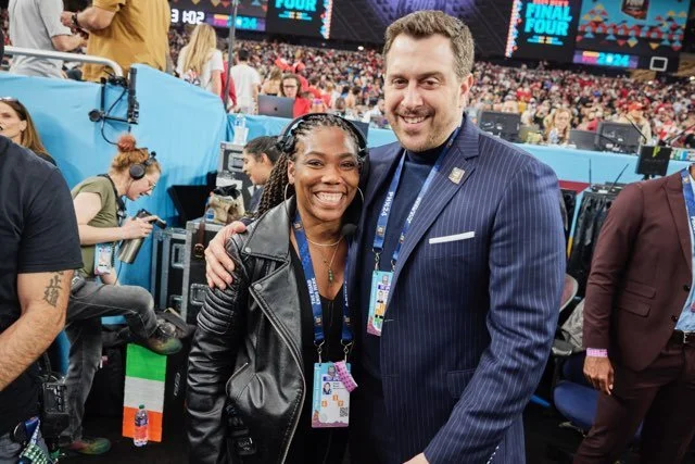 Adam Lefkoe and Nicole Monet pose together for a picture at the NCAA Men's Final Four Tournament in an arena filled with crowd, cameras, and event staff, with digital scoreboard displays in the background.