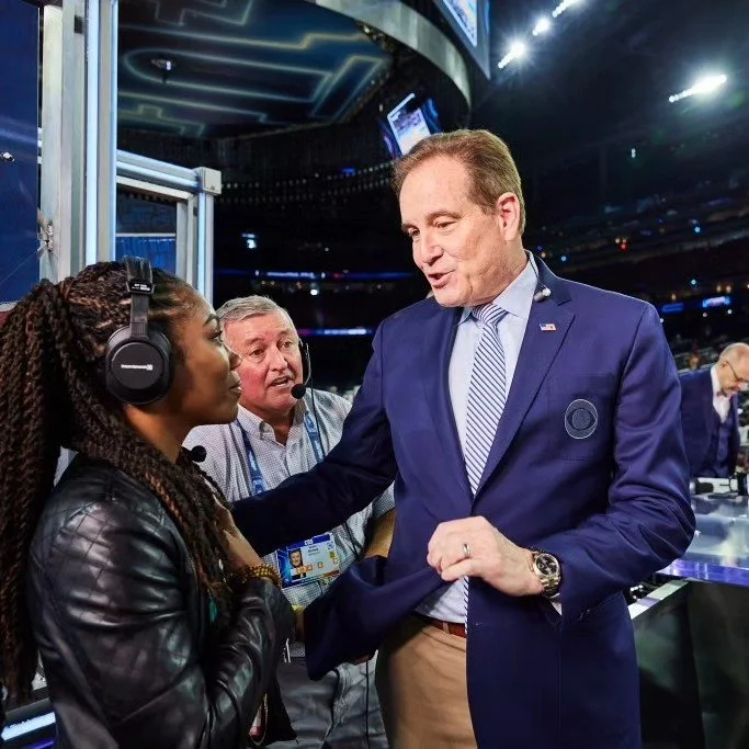 Jim Nantz, sports commentor is complimenting Nicole Monet, who is wearing headphones at an indoor event with dark lighting.