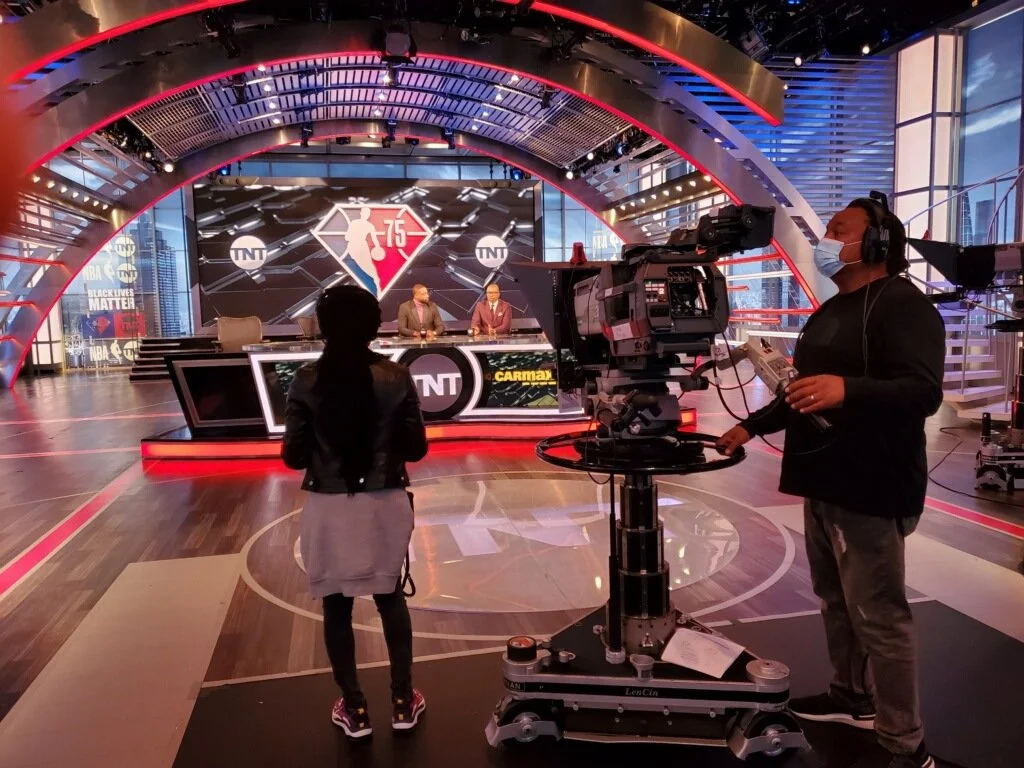 Inside a television studio with a curved red and blue canopy, two sports commentators sit at a desk with the TNT logo and NBA graphics behind them. Two crew members, one with a camera and another with a microphone, prepare to record. The studio featu