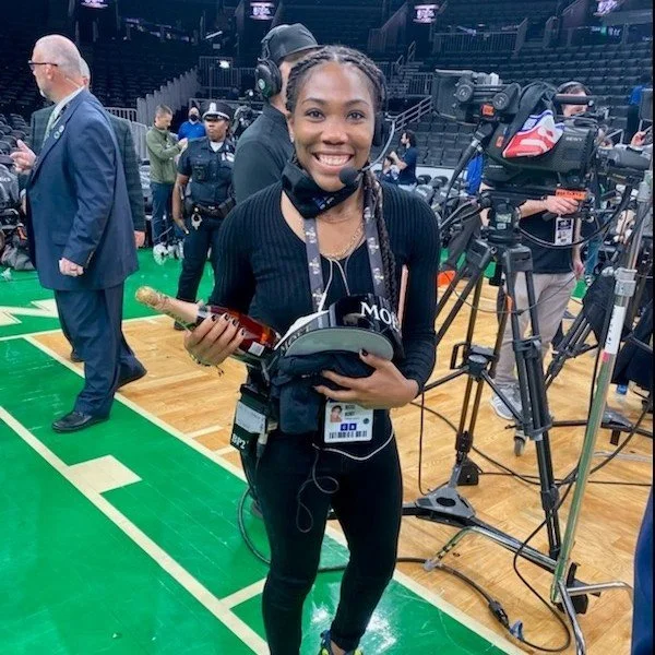 A young woman at a sports event holding a microphone, wearing headphones around her neck, with camera equipment and other crew members in the background.