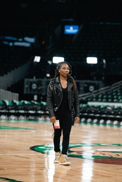 A woman walking alone on a basketball court with empty seats in the background, wearing a black jacket, black pants, tan sneakers, and a headset.