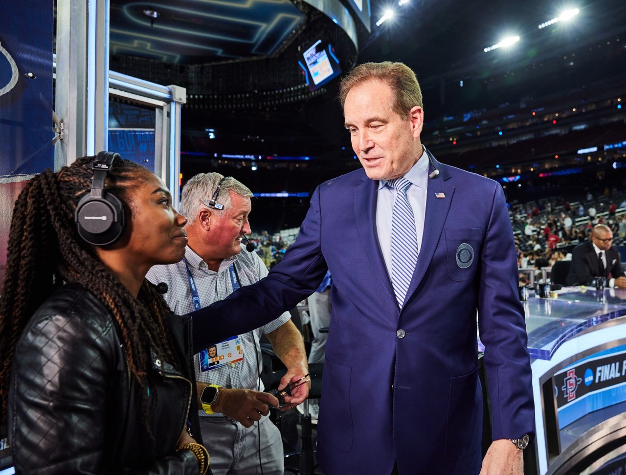 A man in a blue suit shaking hands with a woman wearing headphones at a sports event in a stadium.