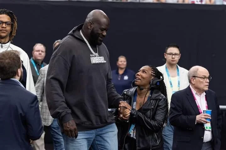 A tall man and a shorter woman are shaking hands and smiling at each other at what appears to be a conference or event. The woman is wearing a black leather jacket and headphones, while the man is dressed casually in a dark hoodie and jeans. Several 