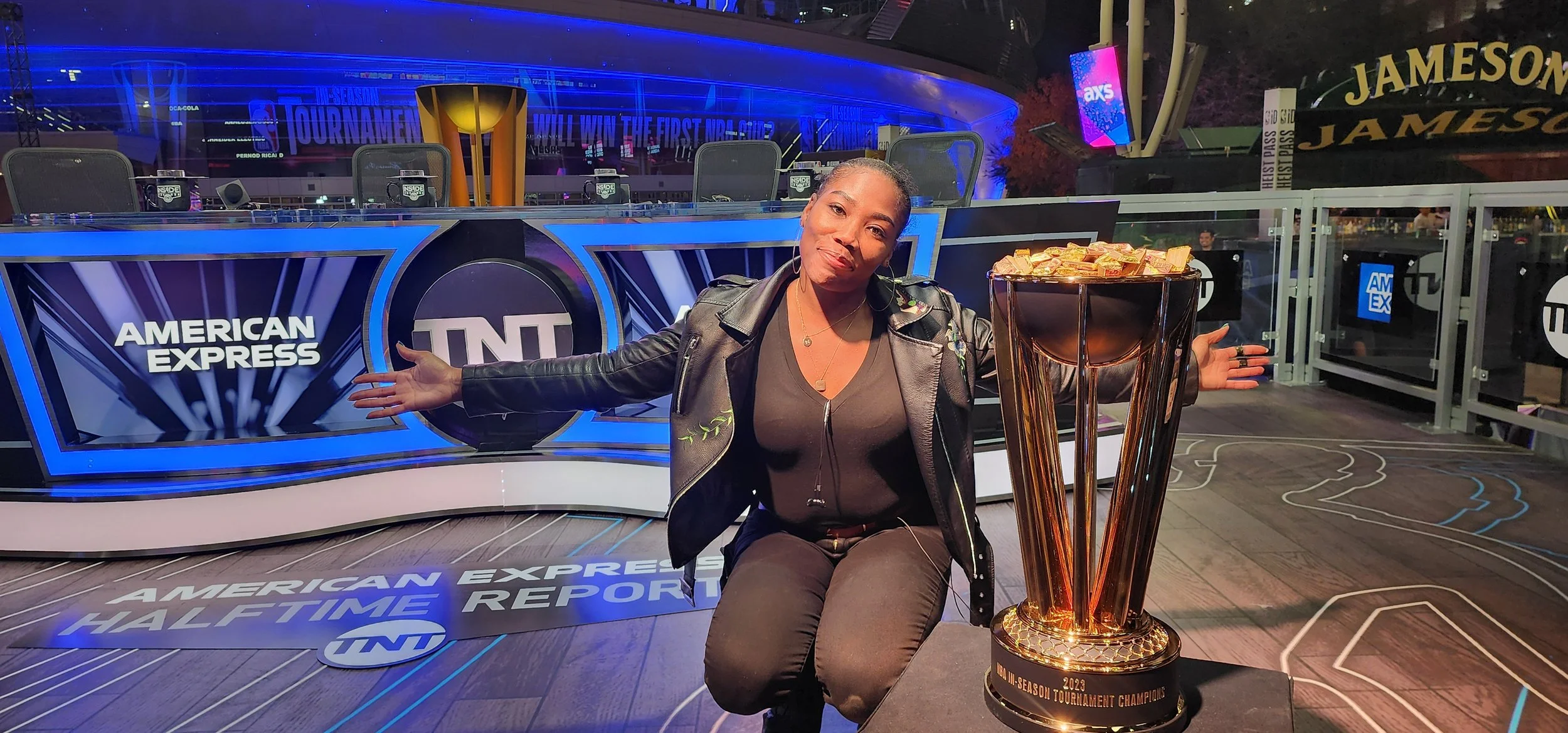 A woman in a black leather jacket and black shirt is sitting in front of a table with America's Cup logo and TNT branding, with her arms open wide. She is smiling and sitting next to a large gold trophy that says '2023 NCAA March Madness Tournament C