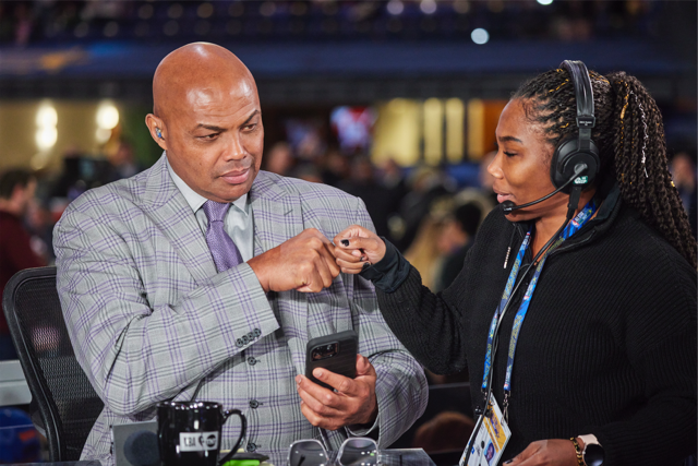 A man in a gray plaid suit looks at his phone while a woman wearing a headset and black jacket shows him something on her wrist at a busy event.