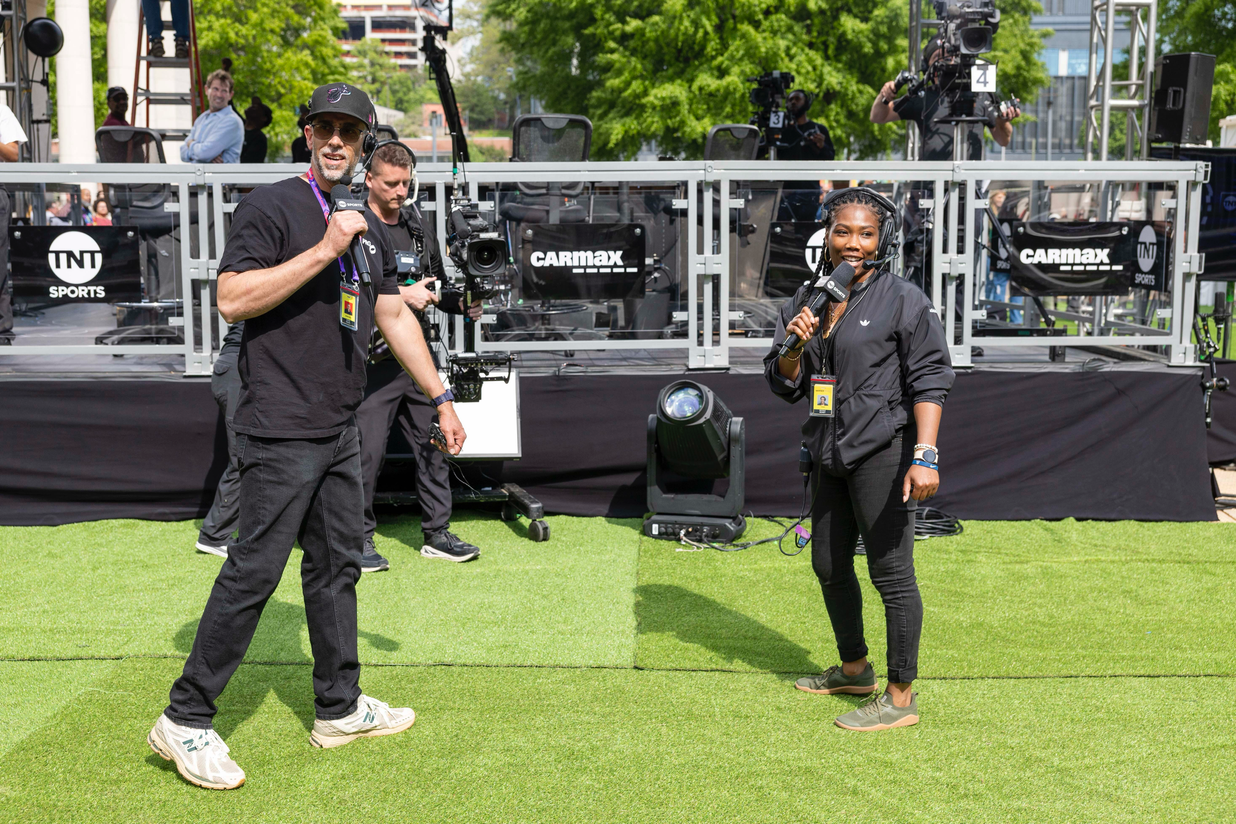 Television sports broadcasters and camera operators setting up at an outdoor event, with a monitor stage and large trees in the background.