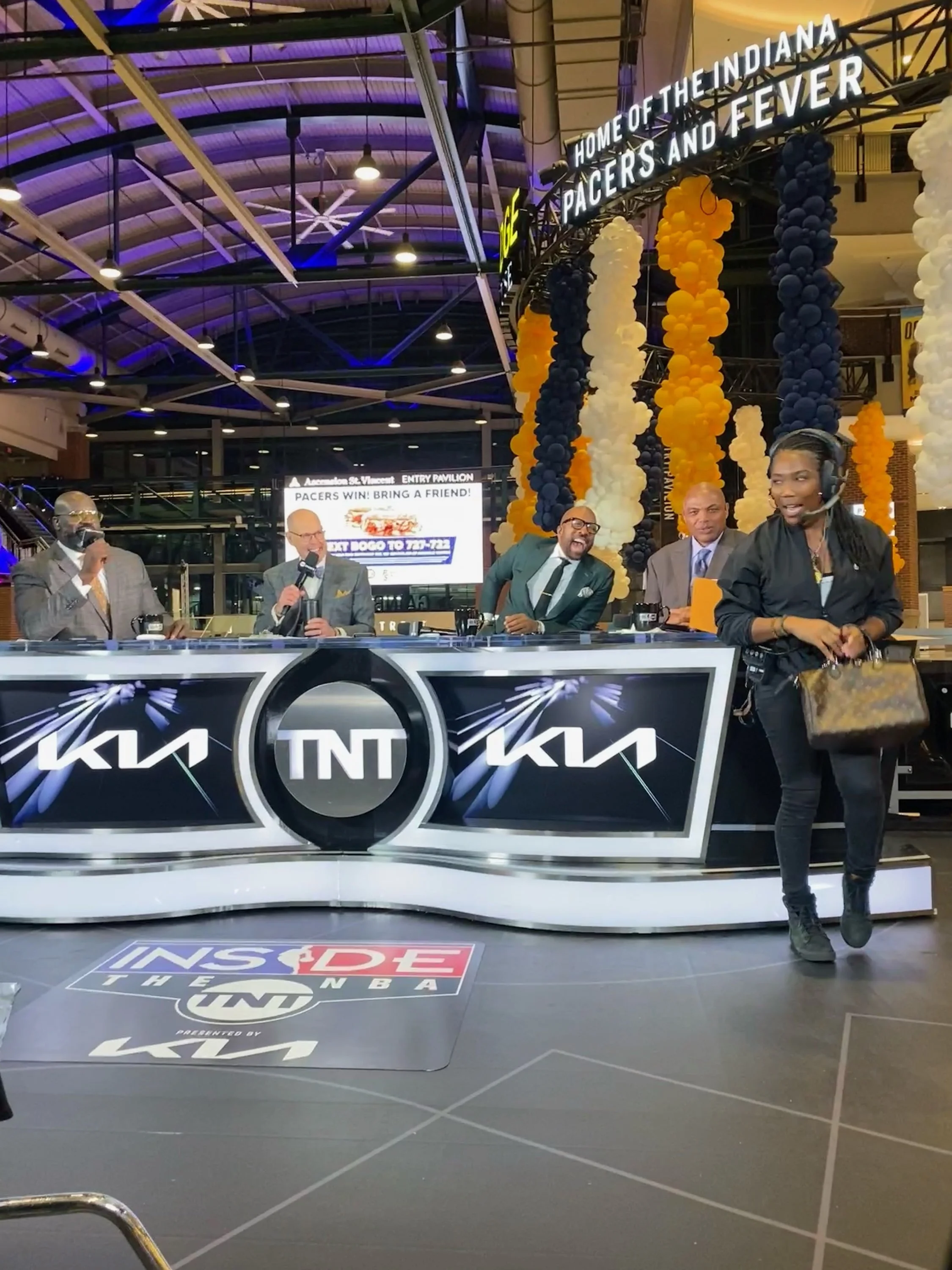 Indoor basketball arena with a broadcast table displaying the TNT and Kia logos, decorated with balloons in blue, white, and orange, celebrating the Indiana Pacers basketball team, with four speakers seated at the table.