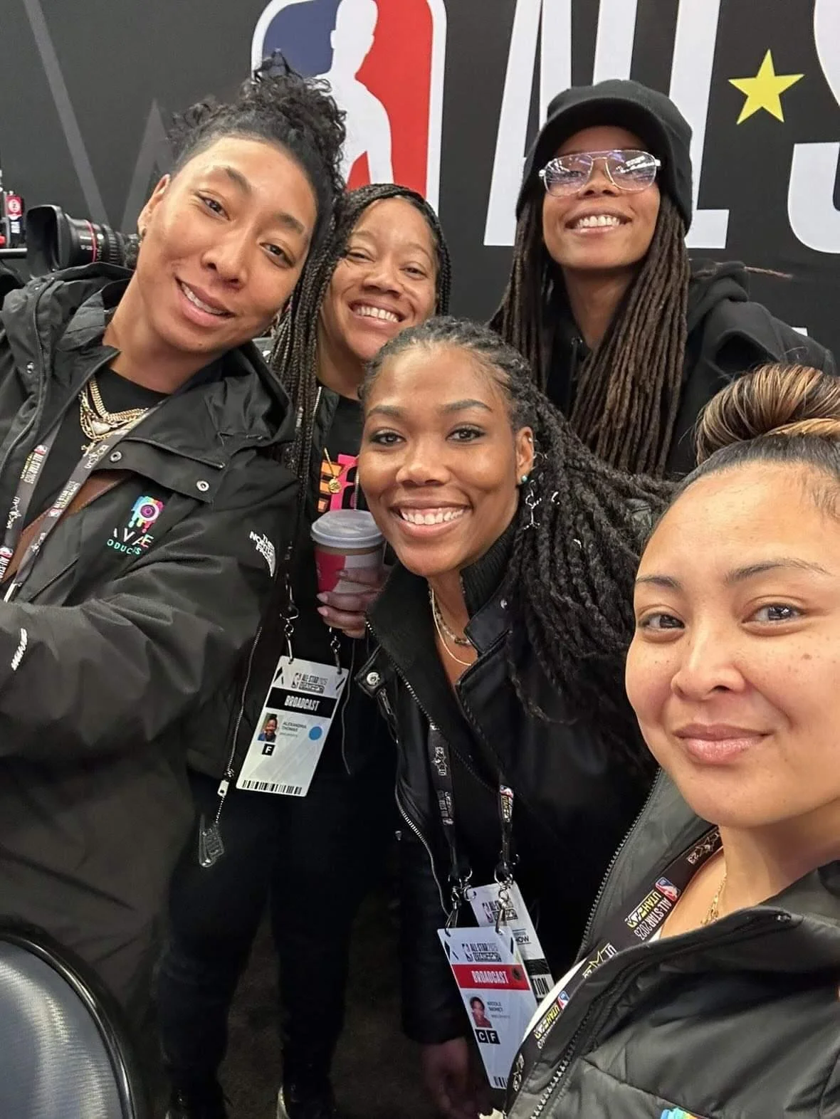 A group of five diverse women smiling and taking a selfie at a conference or event, with event badges and lanyards visible.