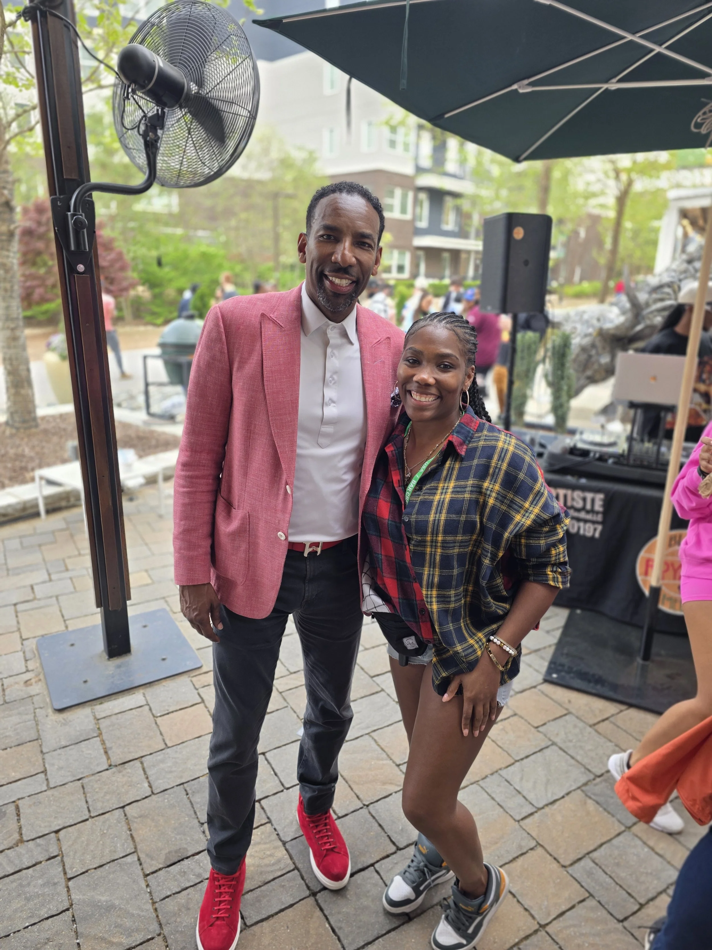 Two people smiling and posing outdoors on a paved area with a fan and a large black speaker in the background. The person on the left is a man wearing a pink blazer, white shirt, dark pants, and red sneakers. The person on the right is a woman wearin