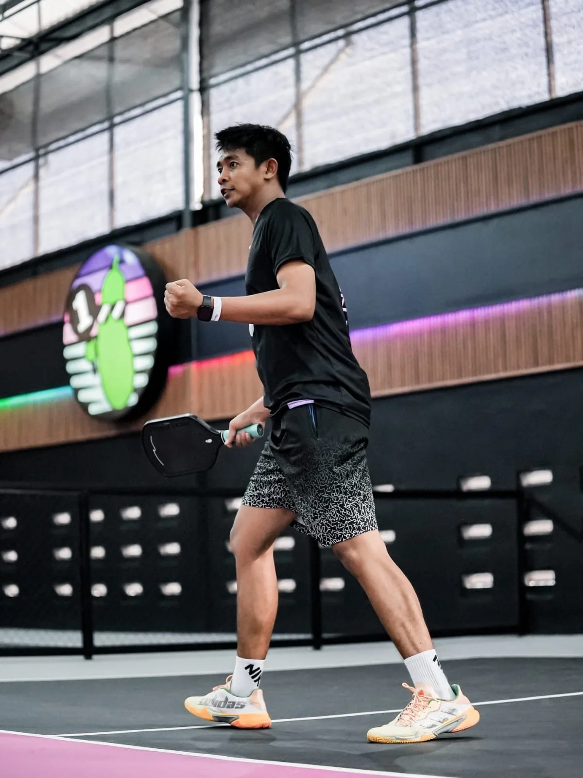 A man playing pickleball on an indoor court, holding a paddle in his right hand, dressed in athletic clothing including a black T-shirt, patterned shorts, white socks, and multicolor sneakers.