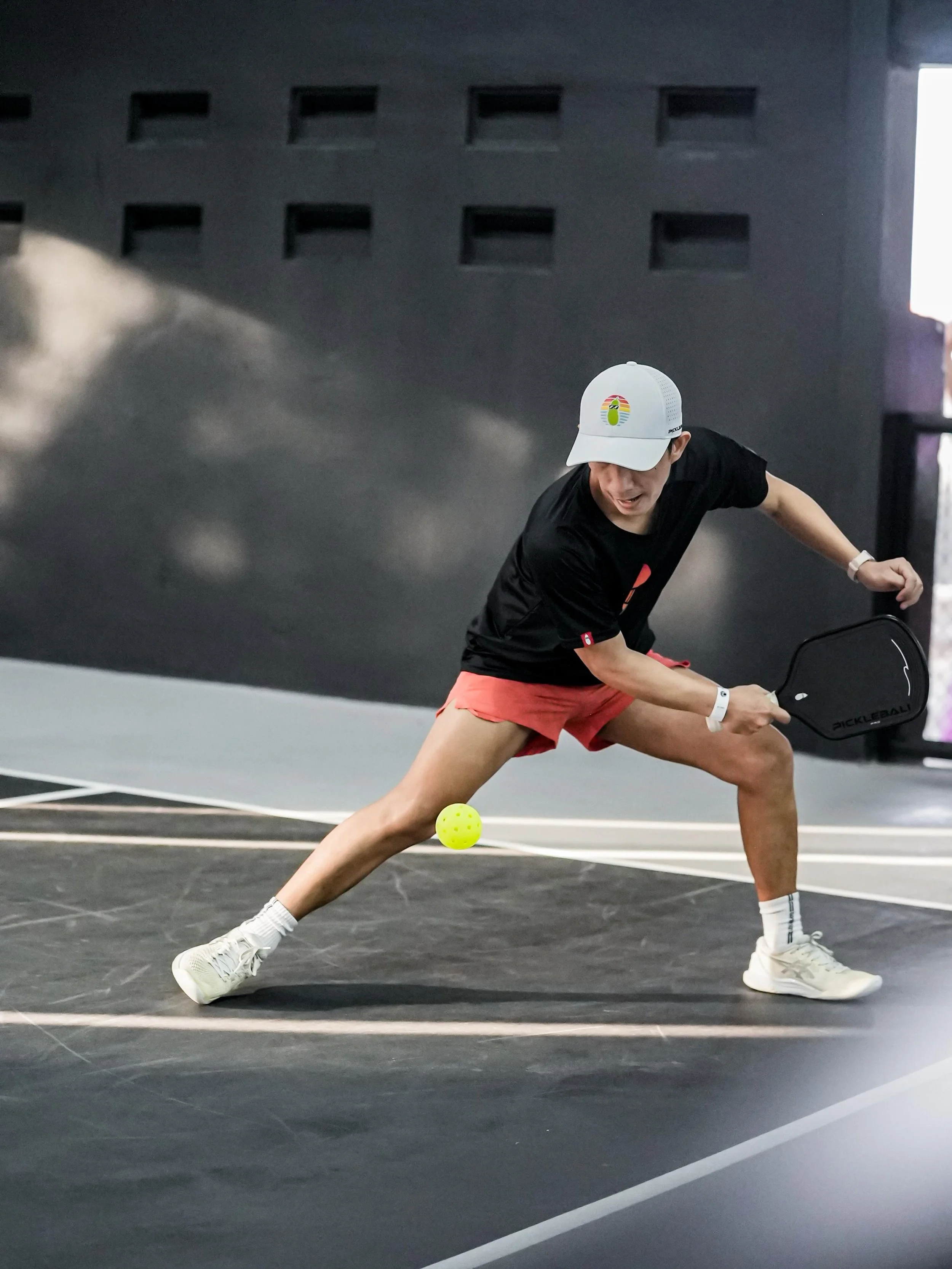 A man playing pickleball on an indoor court, wearing a white cap, black t-shirt, red shorts, and white sneakers, with a yellow pickleball near his racket.