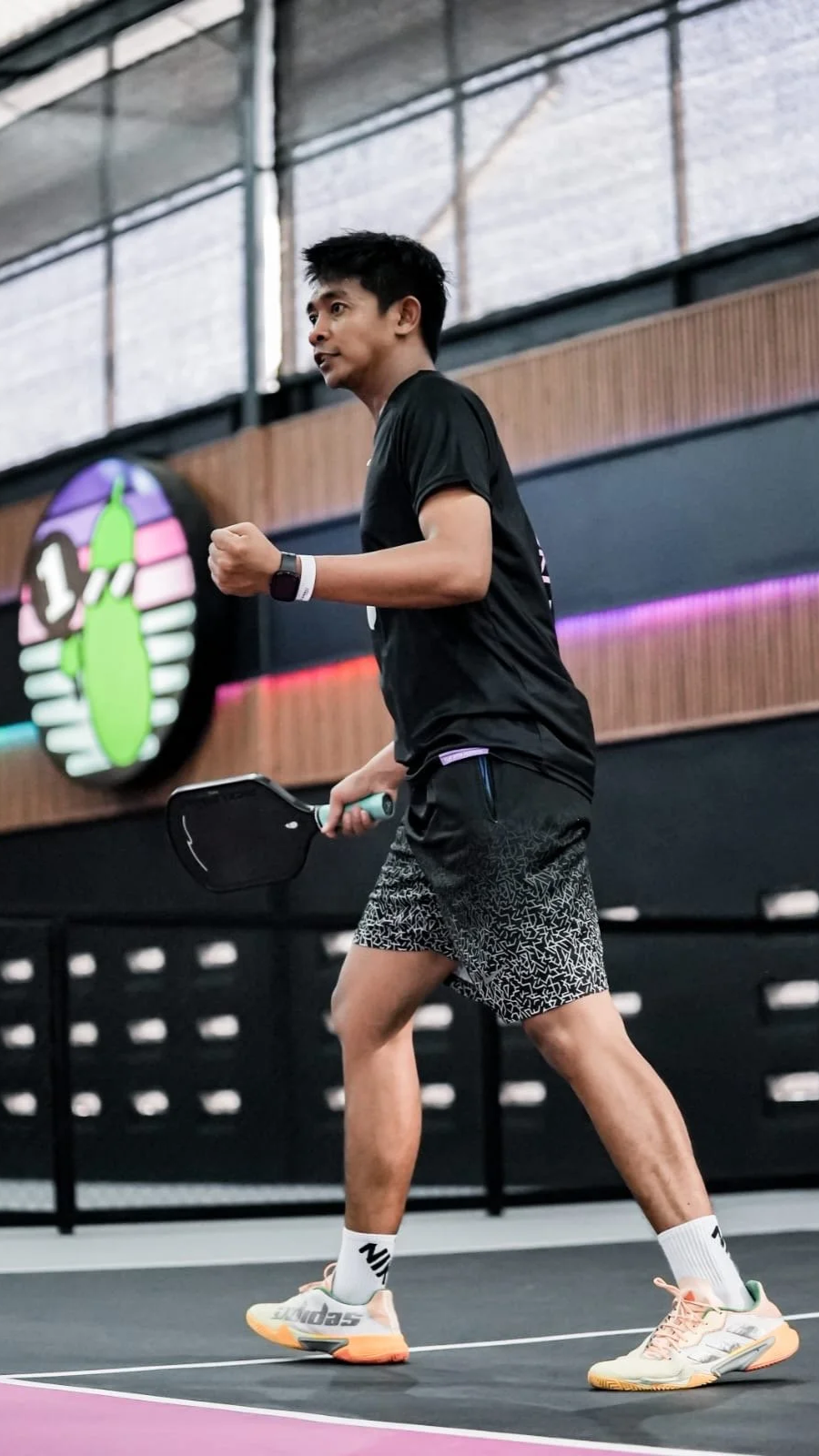 A man in black shorts and a black t-shirt playing pickleball with a paddle on an indoor court.
