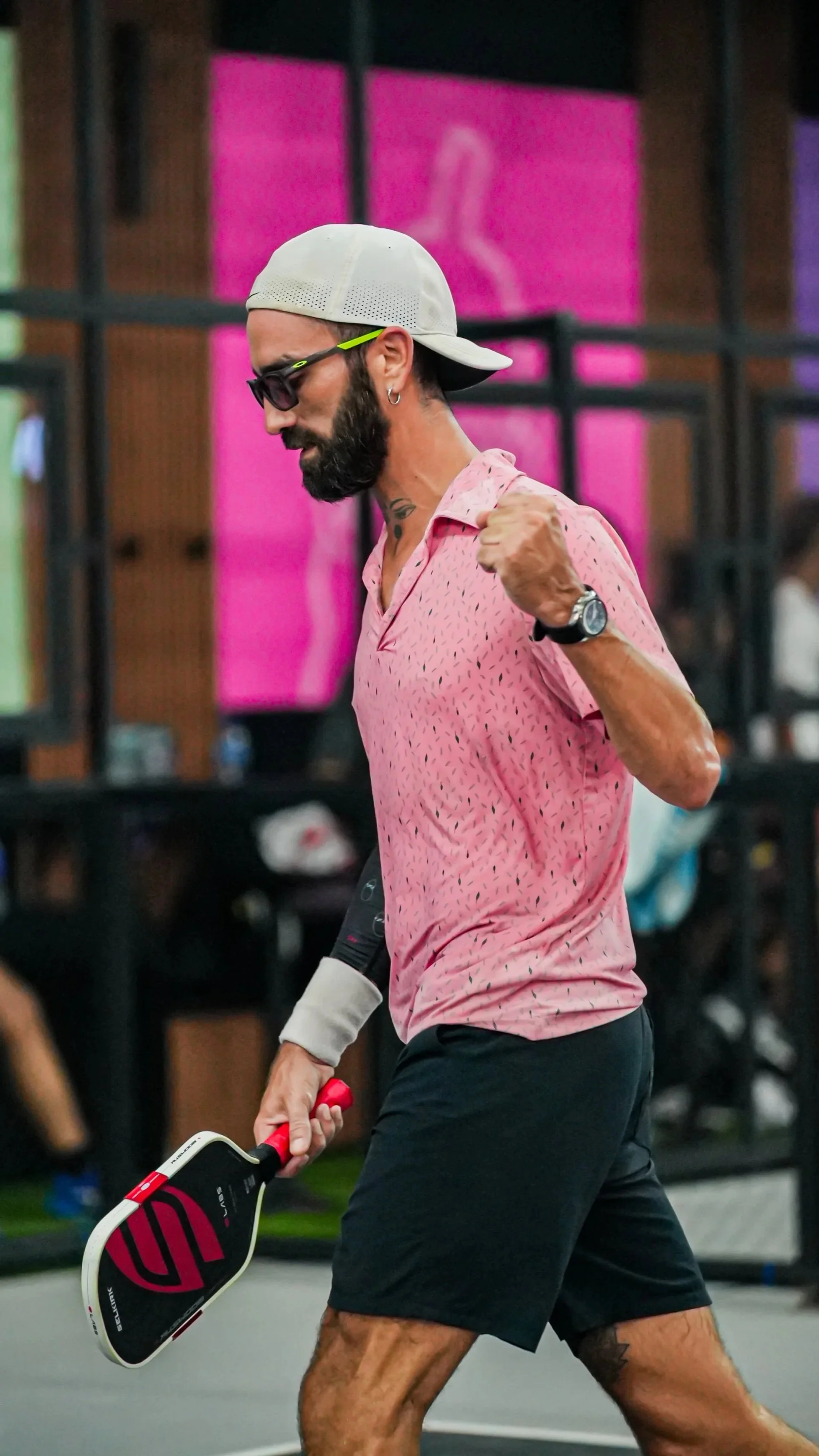 A man with a beard, wearing sunglasses, a backwards cap, a pink patterned shirt, and black shorts, is playing tennis indoors. He has a wristwatch on his left wrist and is holding a tennis racket in his right hand.