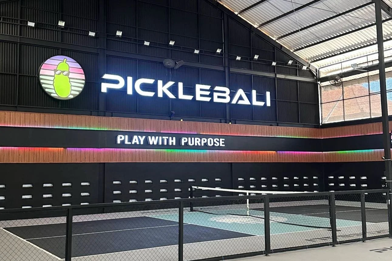 Indoor pickleball court with a large illuminated sign that says 'PICKLEBALL' and a logo of a pickle with sunglasses. Below, a digital sign reads 'PLAY WITH PURPOSE,' and the court is enclosed by a black fence.