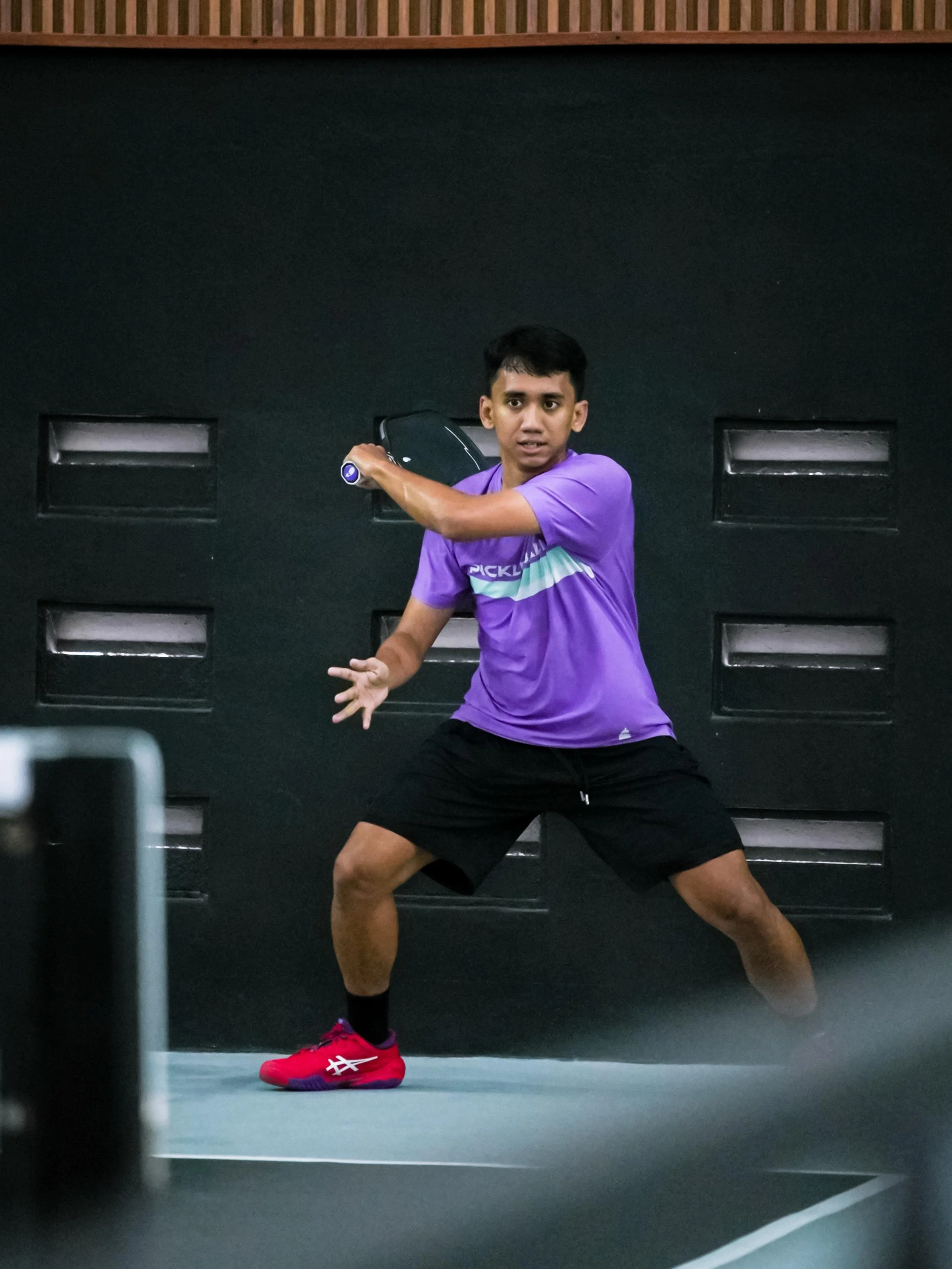A young man playing tennis or pickleball indoors, wearing a purple shirt, black shorts, and red athletic shoes, with a black paddle in his hand, standing in a ready position in front of a black wall with ventilation openings.