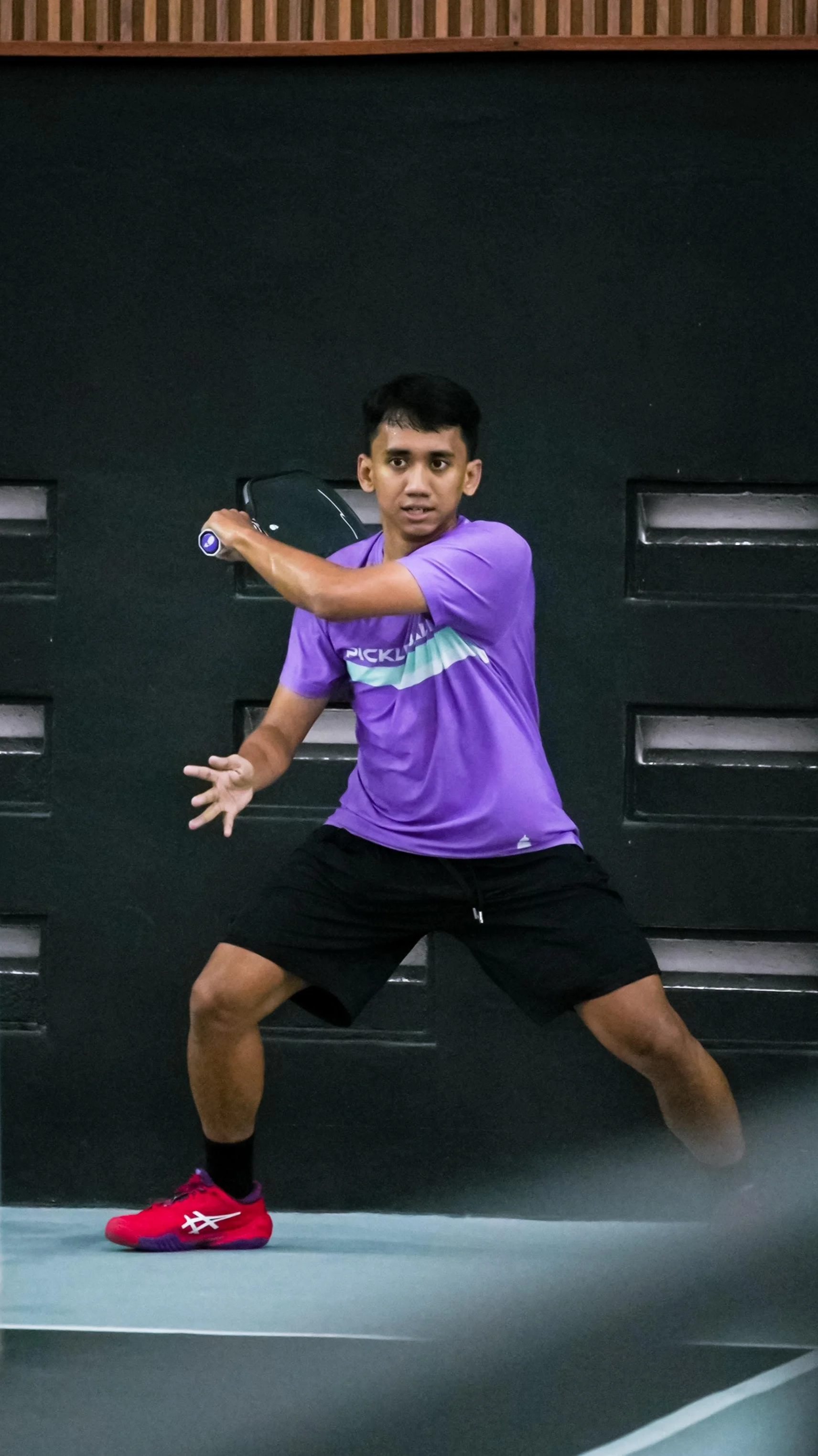A young man in a purple t-shirt and black shorts playing tennis indoors, preparing to hit a shot with a tennis racket.