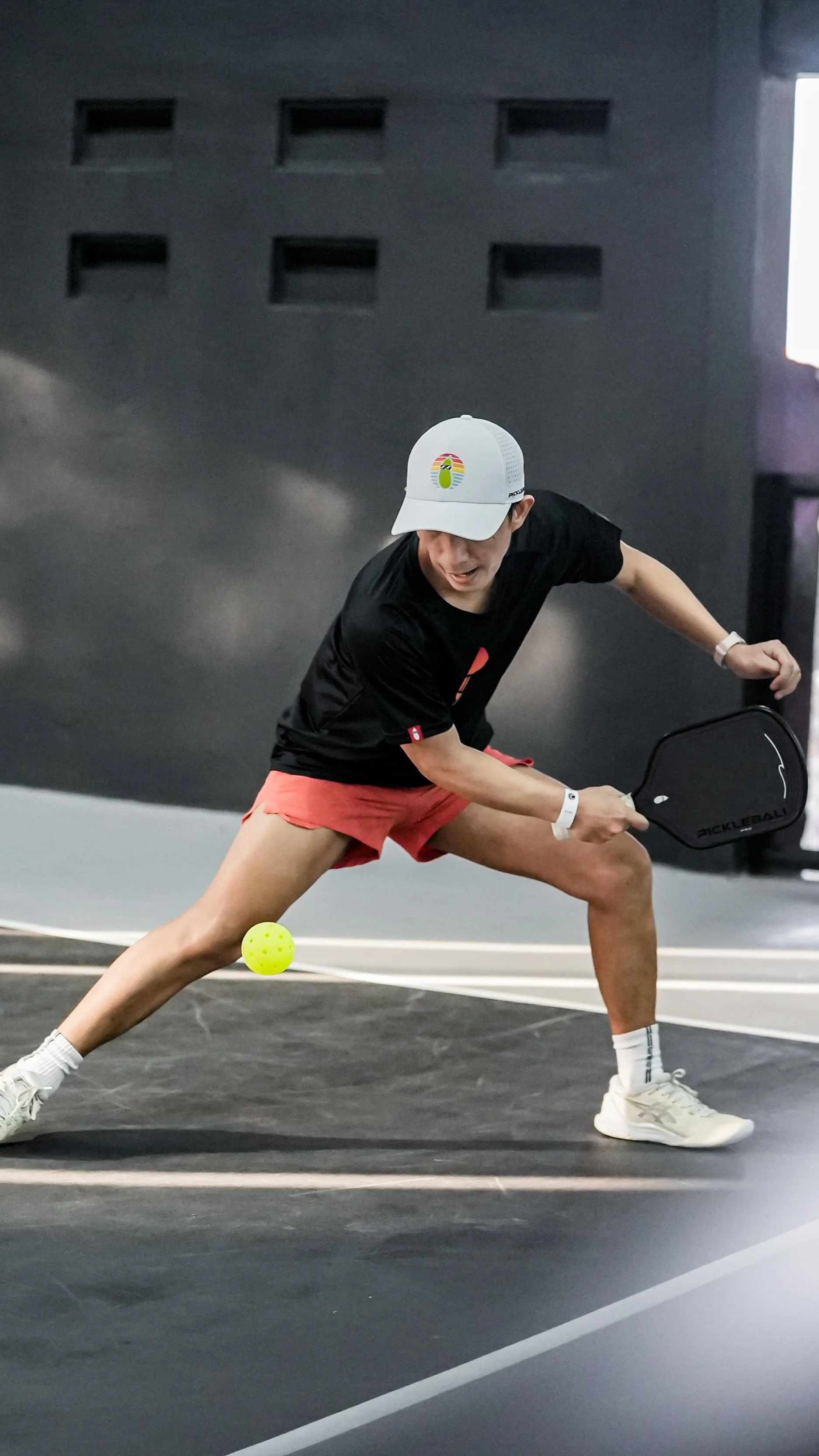 A person playing pickleball on an indoor court, wearing a white cap, black shirt, and pink shorts, with a yellow pickleball nearby.