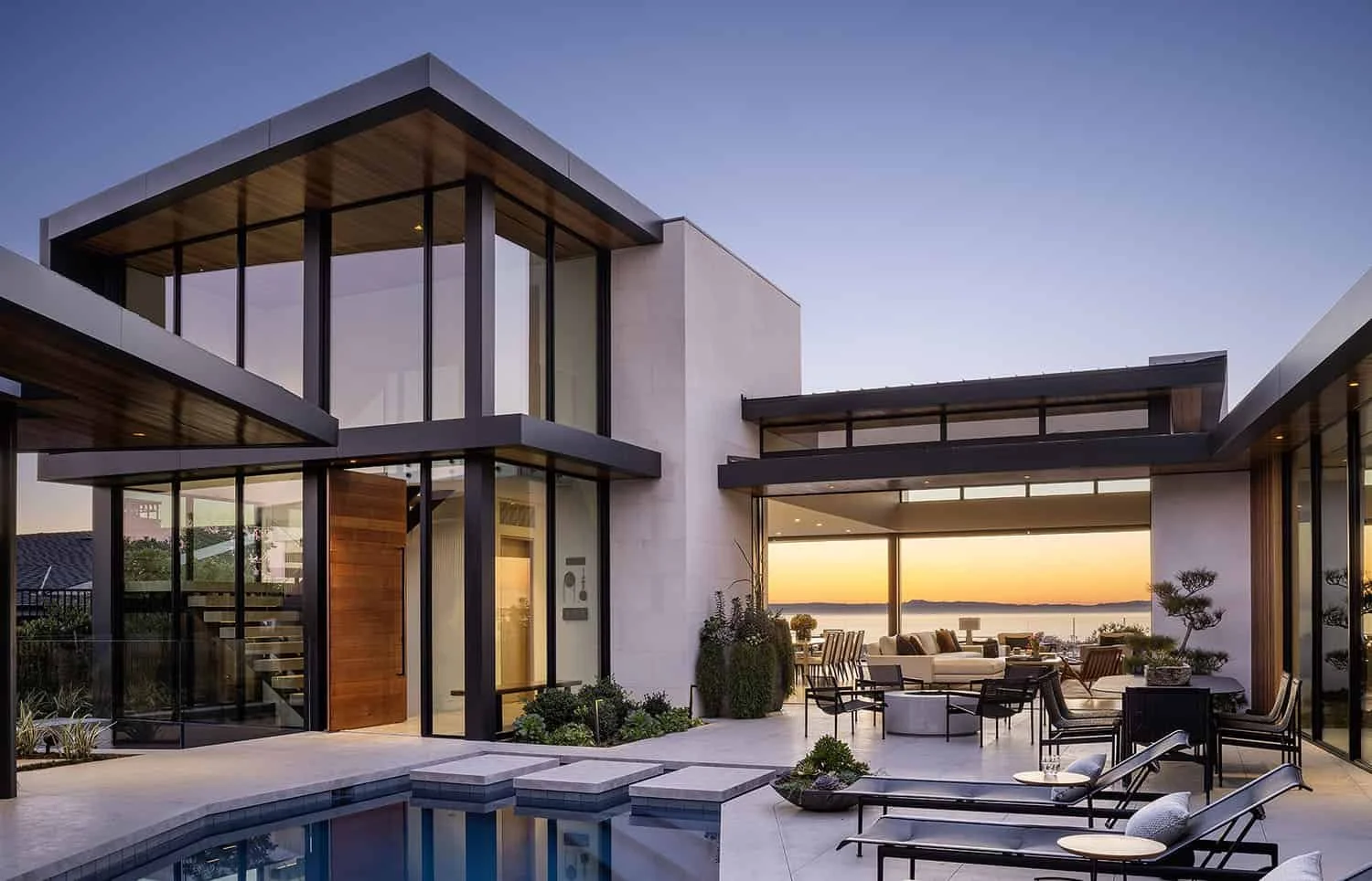 Modern house with large glass windows overlooking water at sunset, outdoor lounge area with chairs and a pool.
