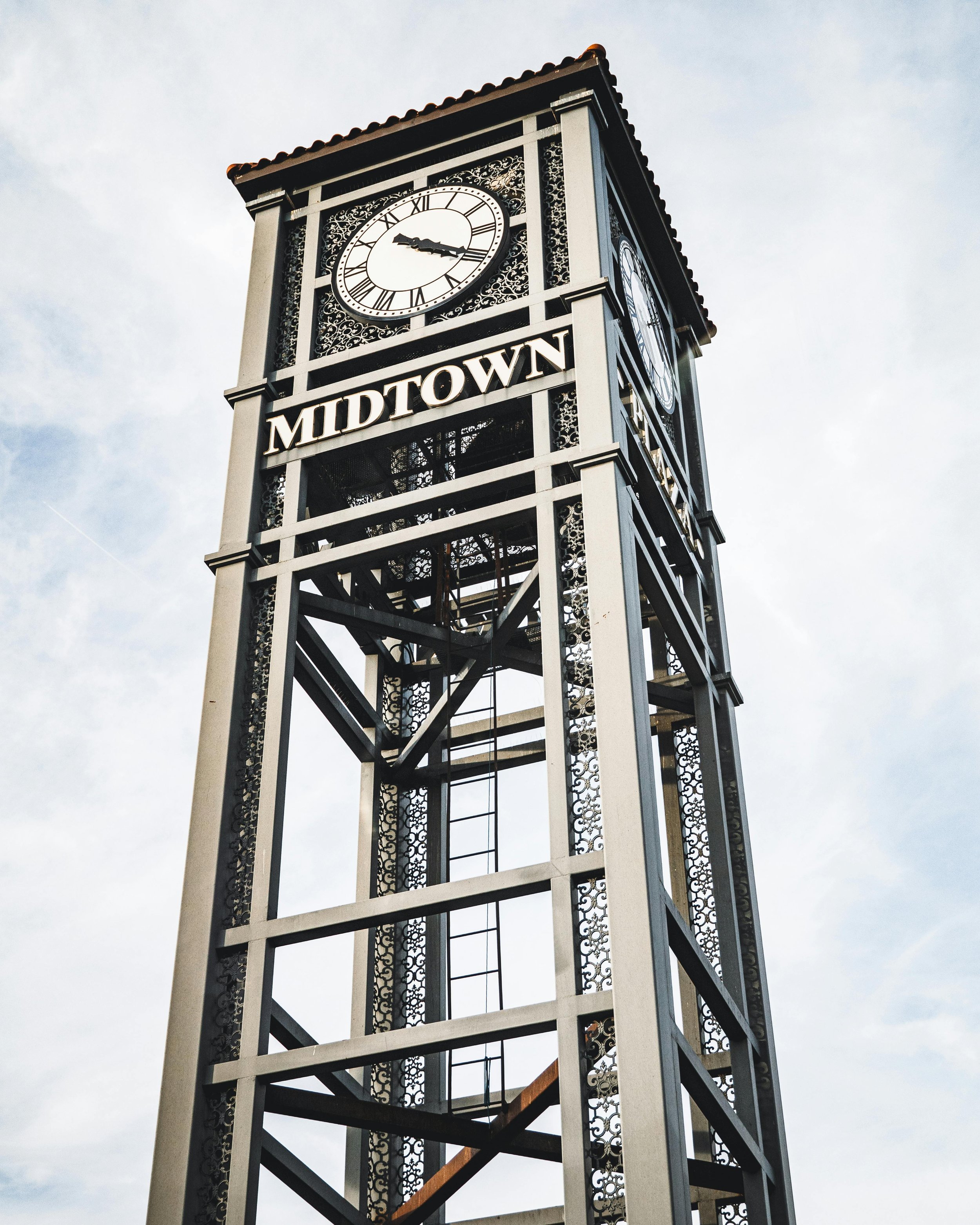 A black and white street clock displaying the time 3:15, with the word "MIDTOWN" below it, set on a decorative metal framework against a cloudy sky.