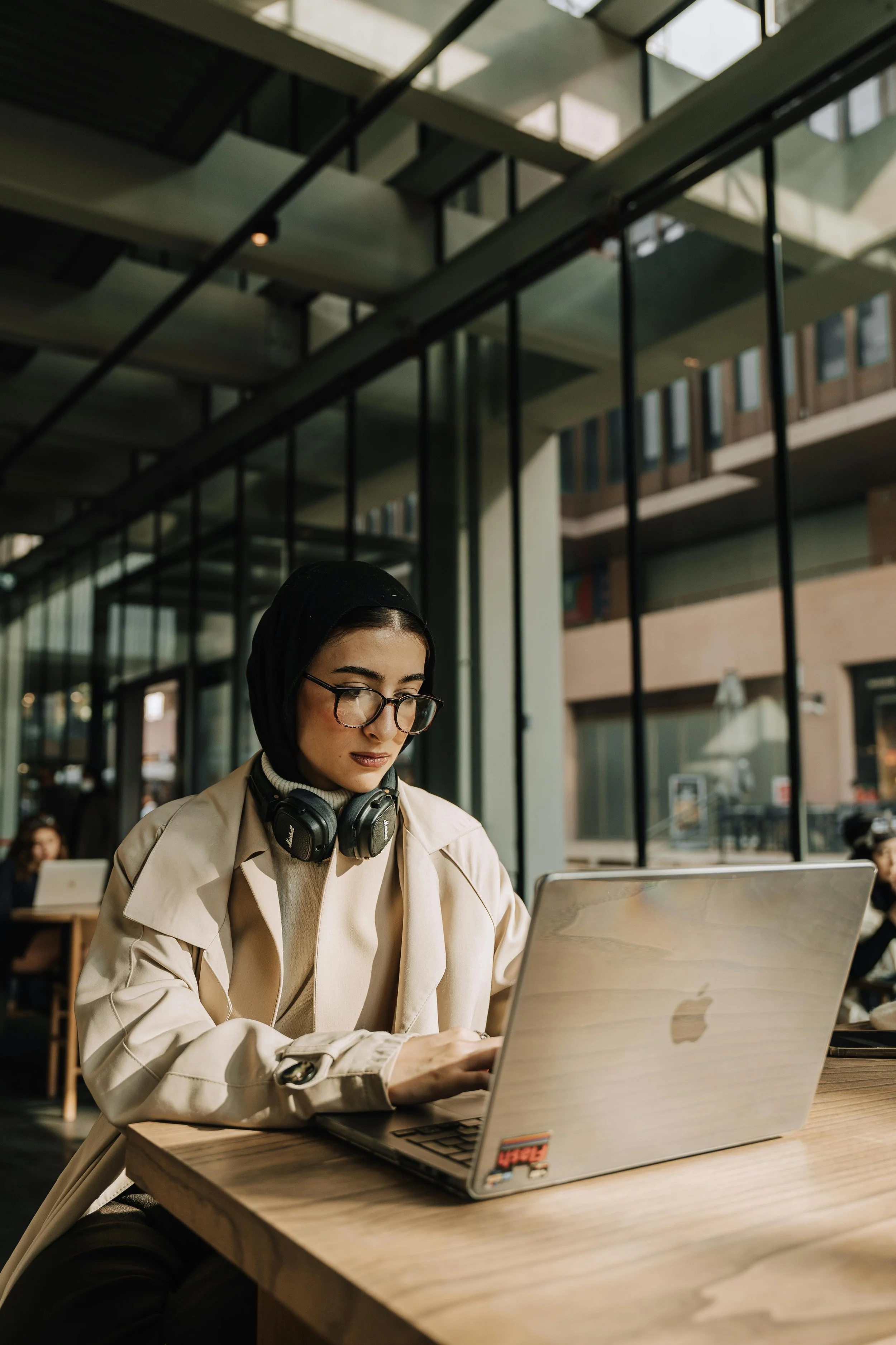 A woman with glasses and a black headscarf working on a silver MacBook laptop at a wooden table inside a modern, glass-walled building with multiple levels.
