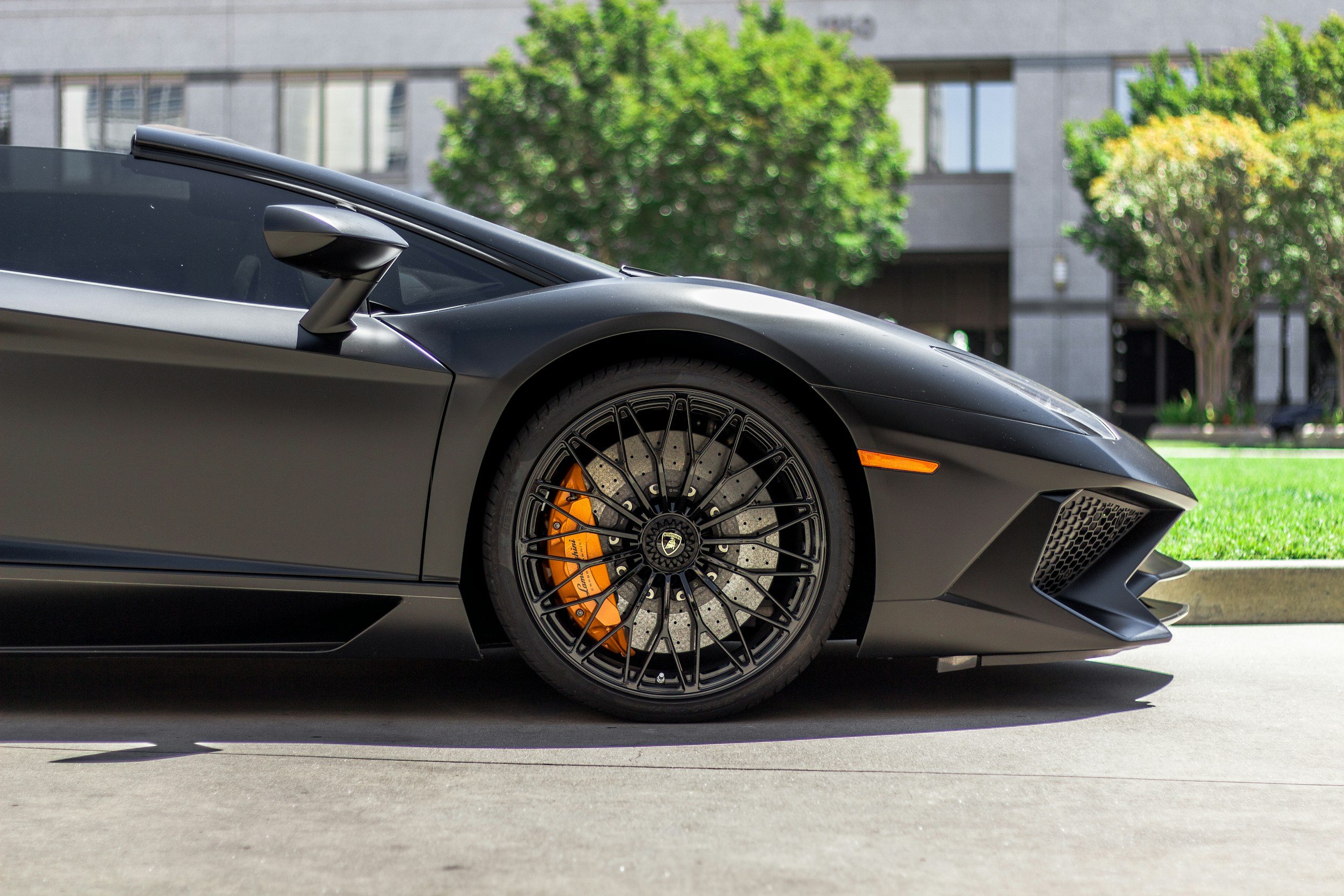 Close-up of the front side of a matte gray Lamborghini Huracan with black wheels and orange brake calipers, parked on a street with a modern building and trees in the background.