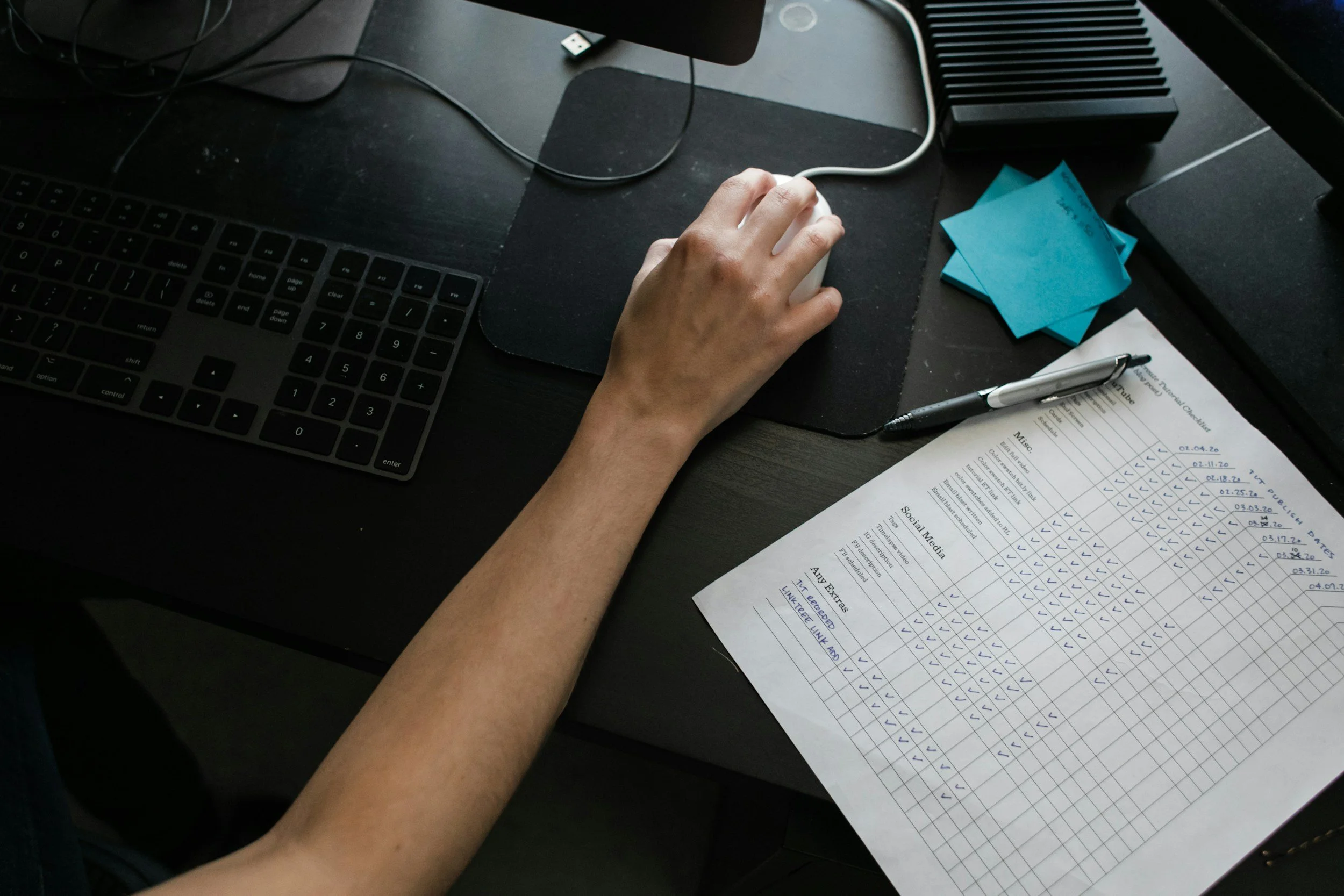 A person using a computer mouse on a black desk with a keyboard, a sheet of paper with a chart labeled 'Social Media,' sticky notes, a pen, and electronic devices.