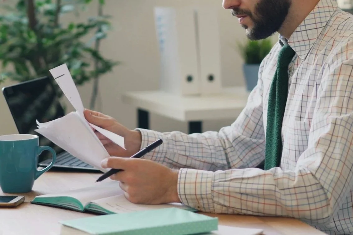 A man in a button-up shirt and green tie sitting at a desk, reviewing papers with a pen in hand, office setting with mug, open notebook, and computer.