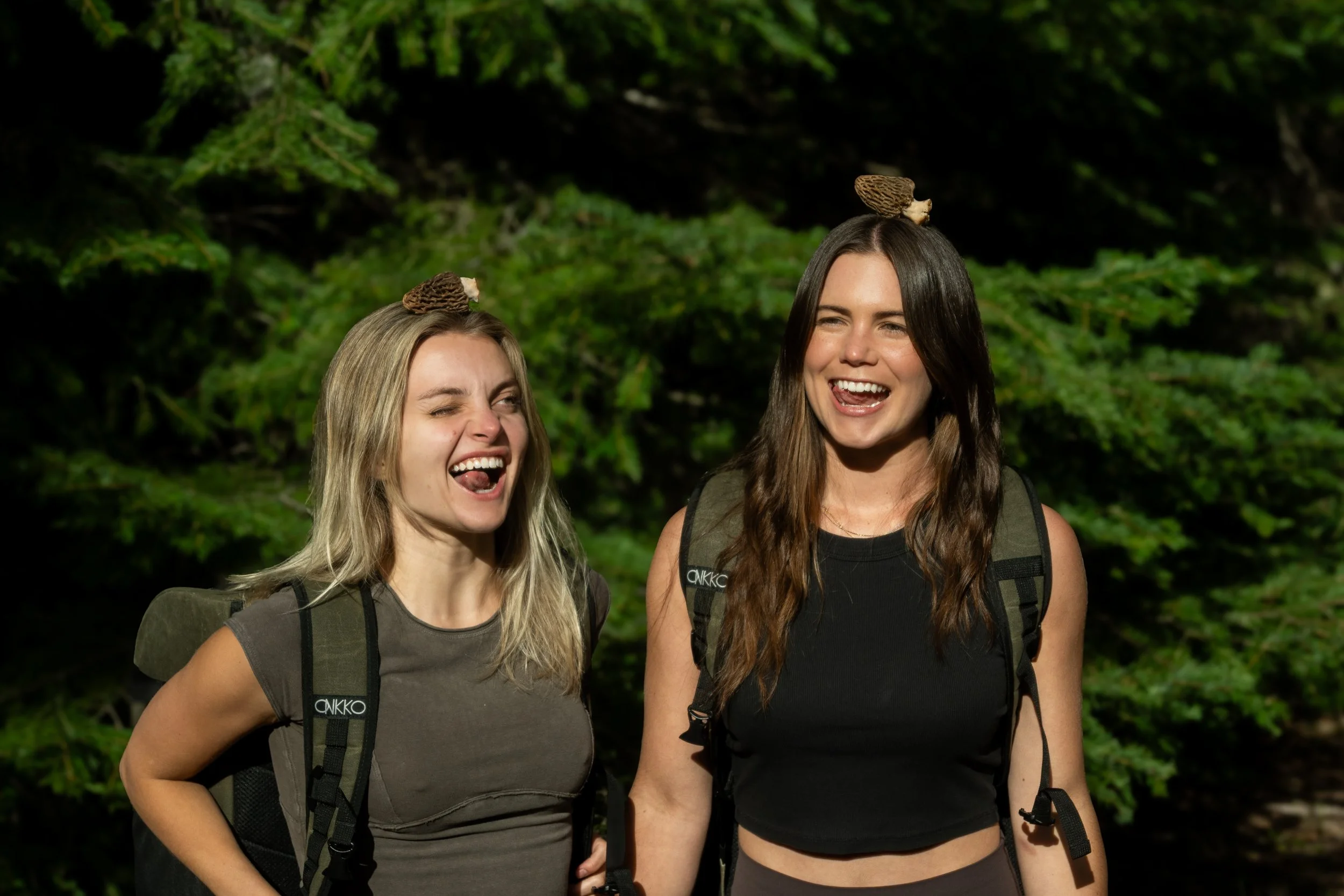Two young women outdoors in a forest, smiling and sticking out their tongues, with morel mushrooms on their heads, wearing foraging backpacks and casual clothing.