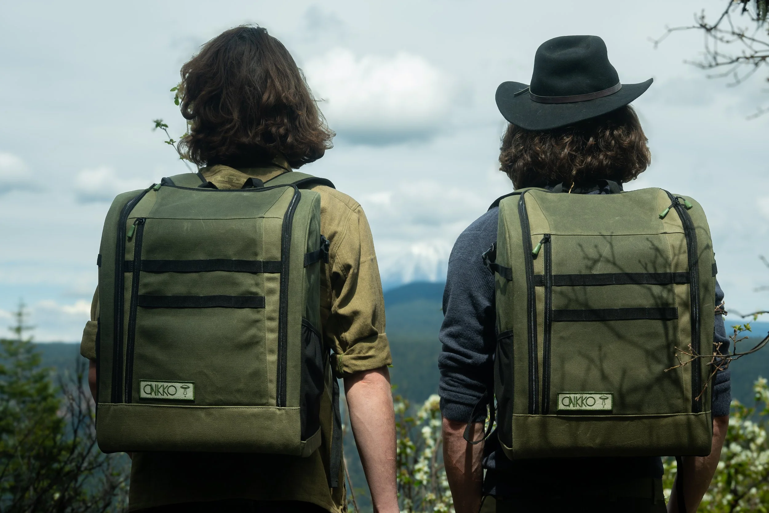 Two men with mushroom foraging backpacks walking outdoors, and looking at a scenic landscape with mountains and sky.