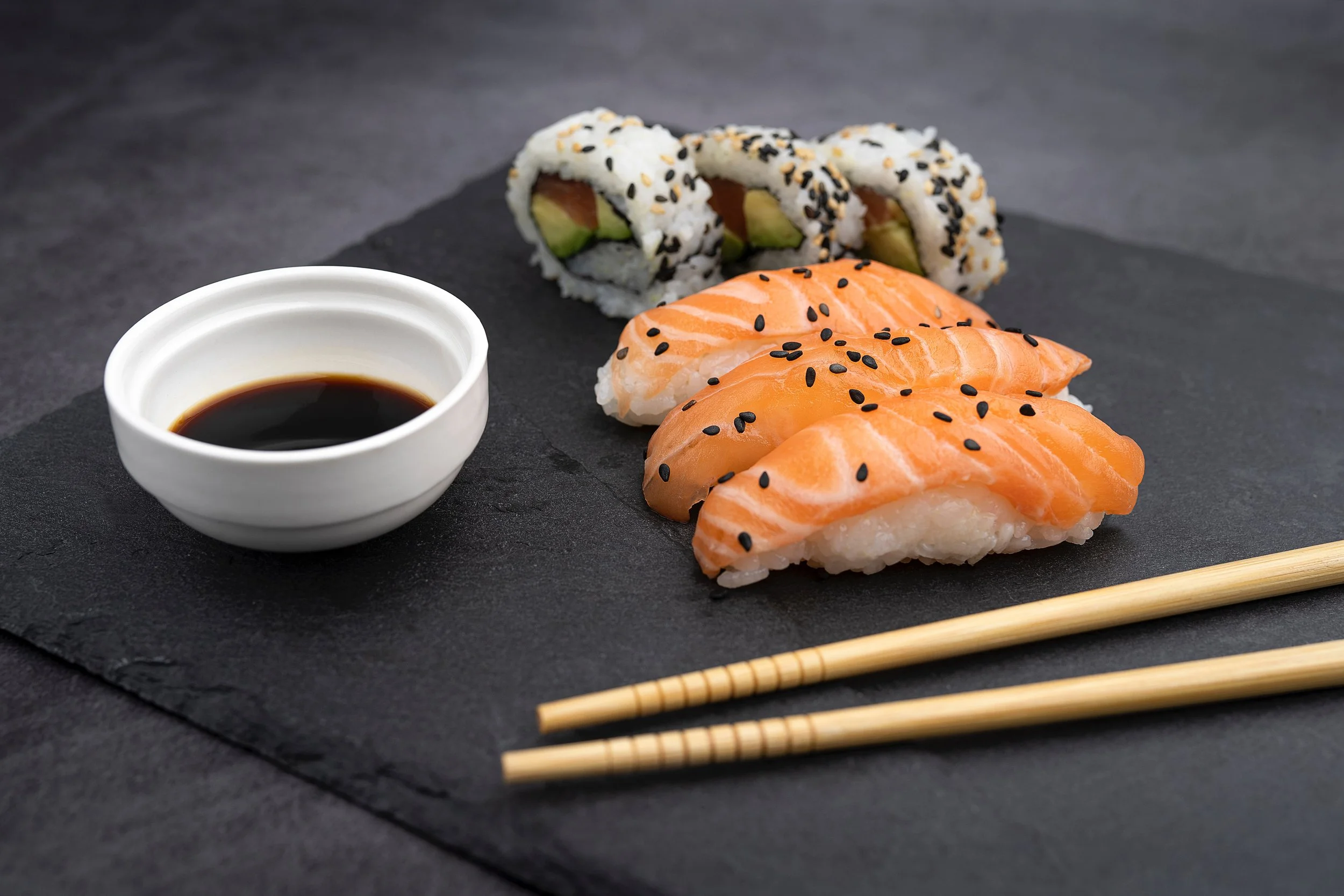 Sushi rolls with sesame seeds, sashimi with black sesame seeds, soy sauce in a small white bowl, and light-colored chopsticks on a black slate surface.