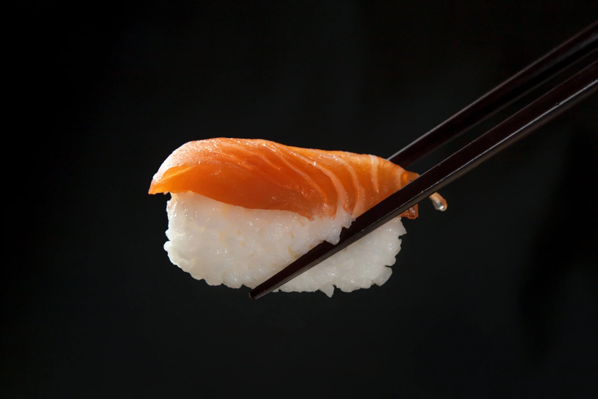 Close-up of a piece of salmon nigiri sushi with rice at the bottom, held by chopsticks against a black background.