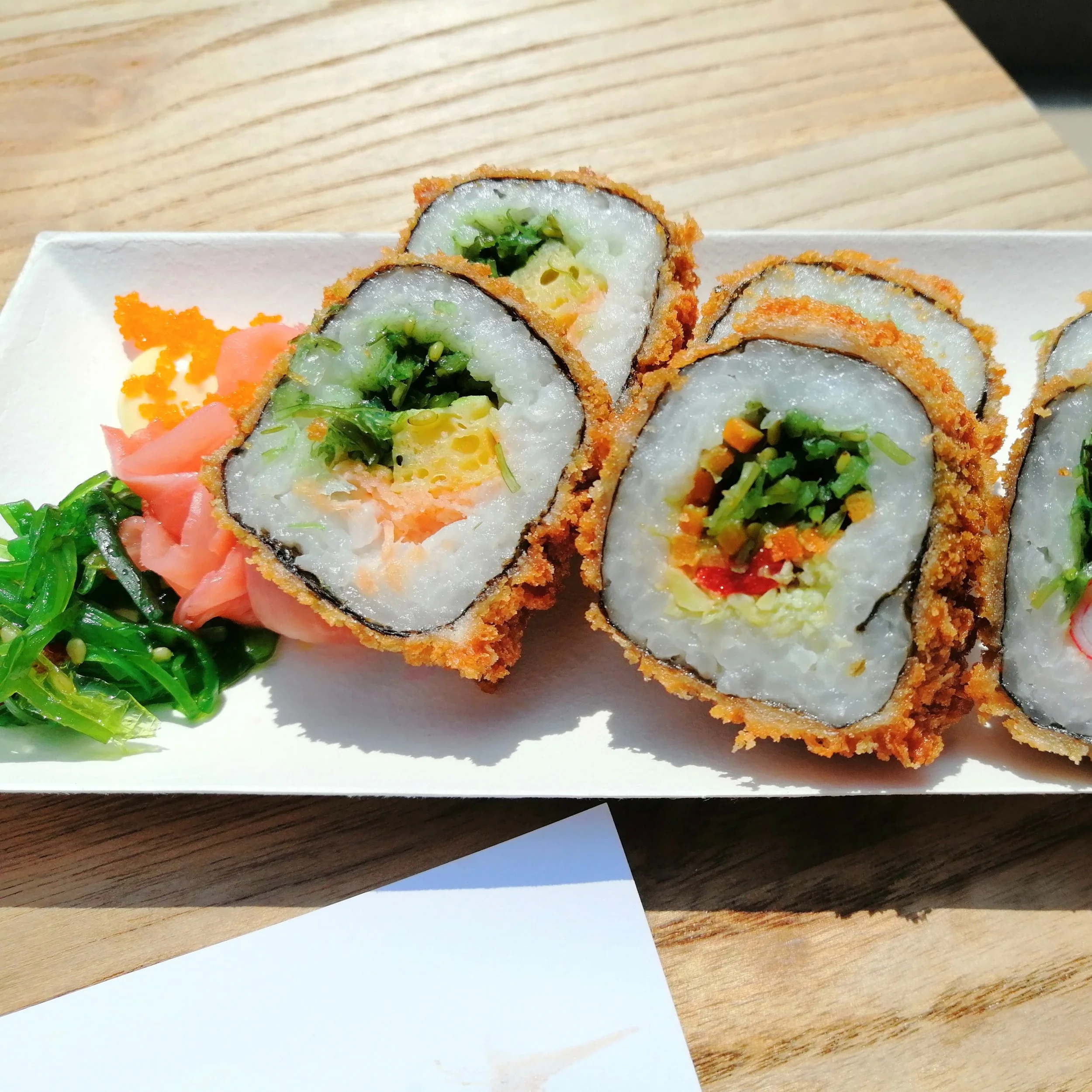 Close-up of several slices of sushi rolls on a white plate, with green seaweed salad and pickled ginger garnishes, placed on a wooden surface.