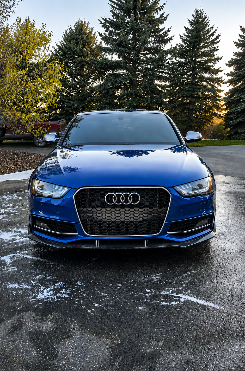 A blue Audi sedan parked on a wet driveway with trees in the background during sunset.