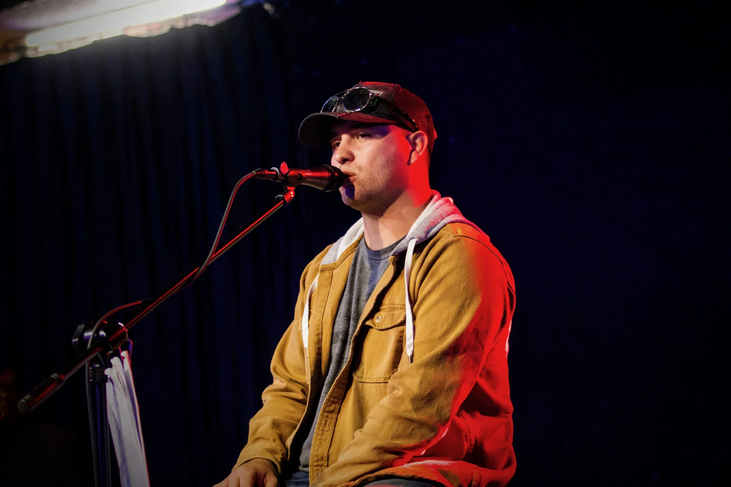 A man performing music on stage, singing into a microphone, wearing a red cap, sunglasses on his head, and a mustard-colored jacket.