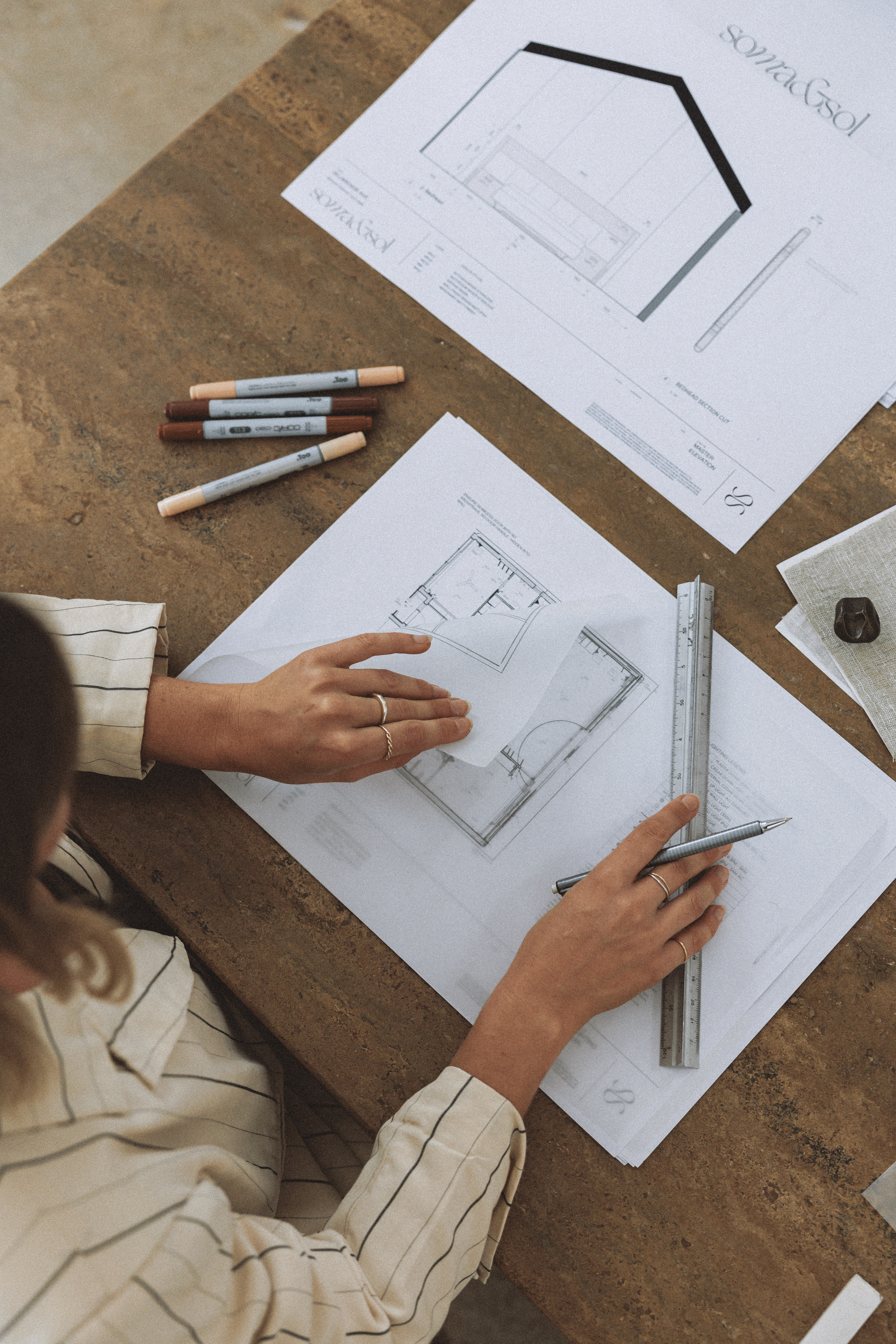 Architectural blueprints on a wooden table with markers, a ruler, and fabric samples, with a person's hand pointing at a drawing.
