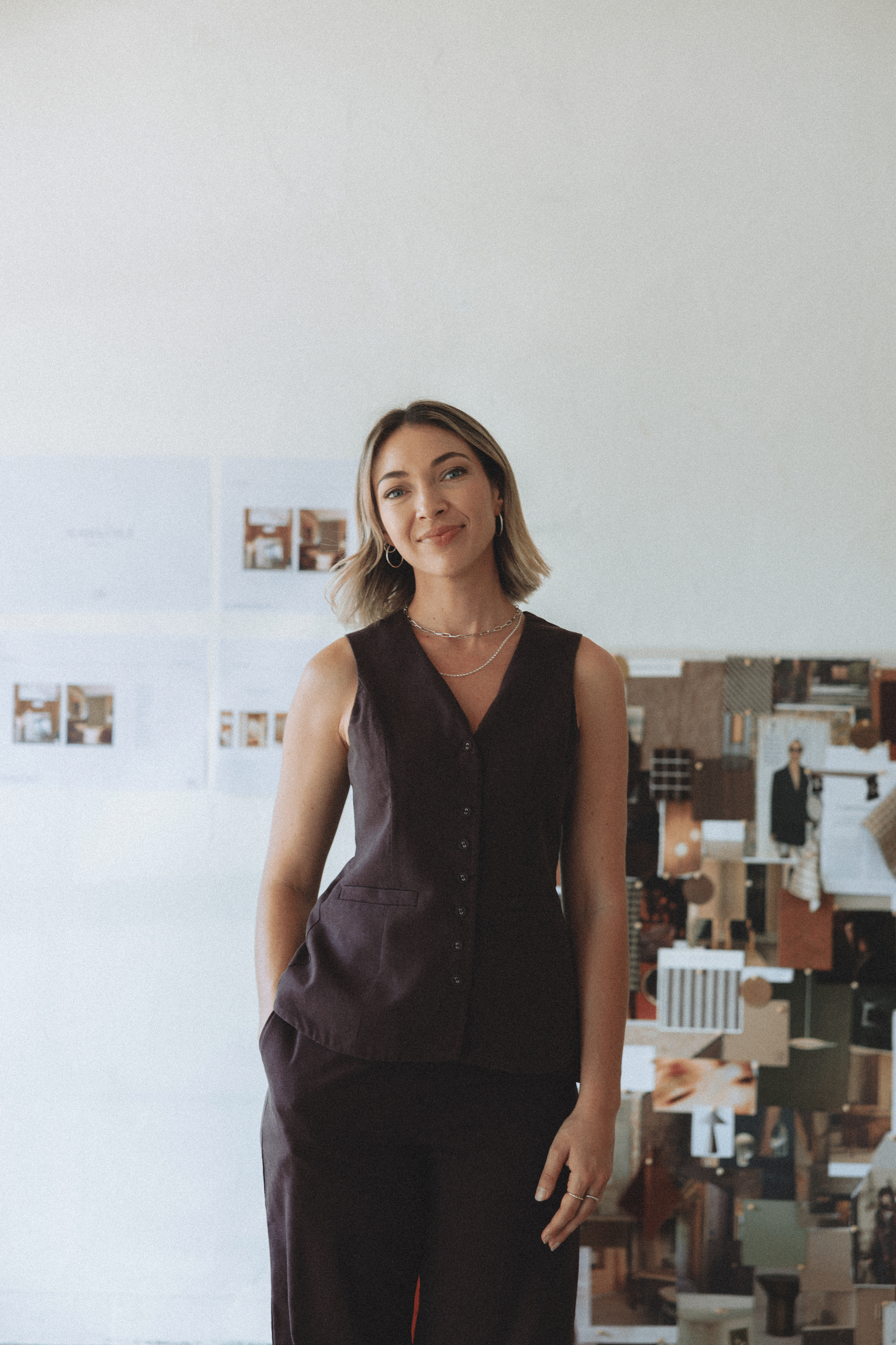 A woman with shoulder-length blonde hair and hoop earrings, smiling gently, wearing a dark sleeveless buttoned dress, standing in an art gallery or studio with artwork on the walls and shelves visible in the background.