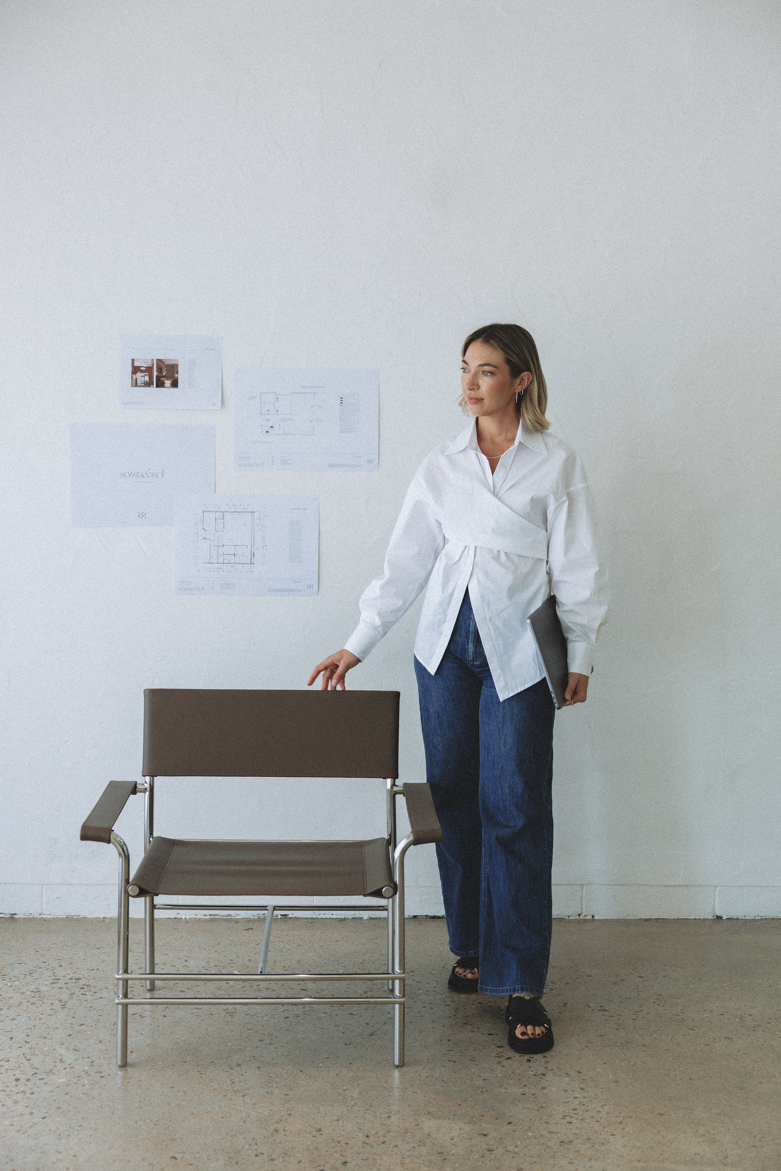 Woman standing in a room with architectural blueprints on the wall, holding a black folder, and reaching for a tan chair in front of her.