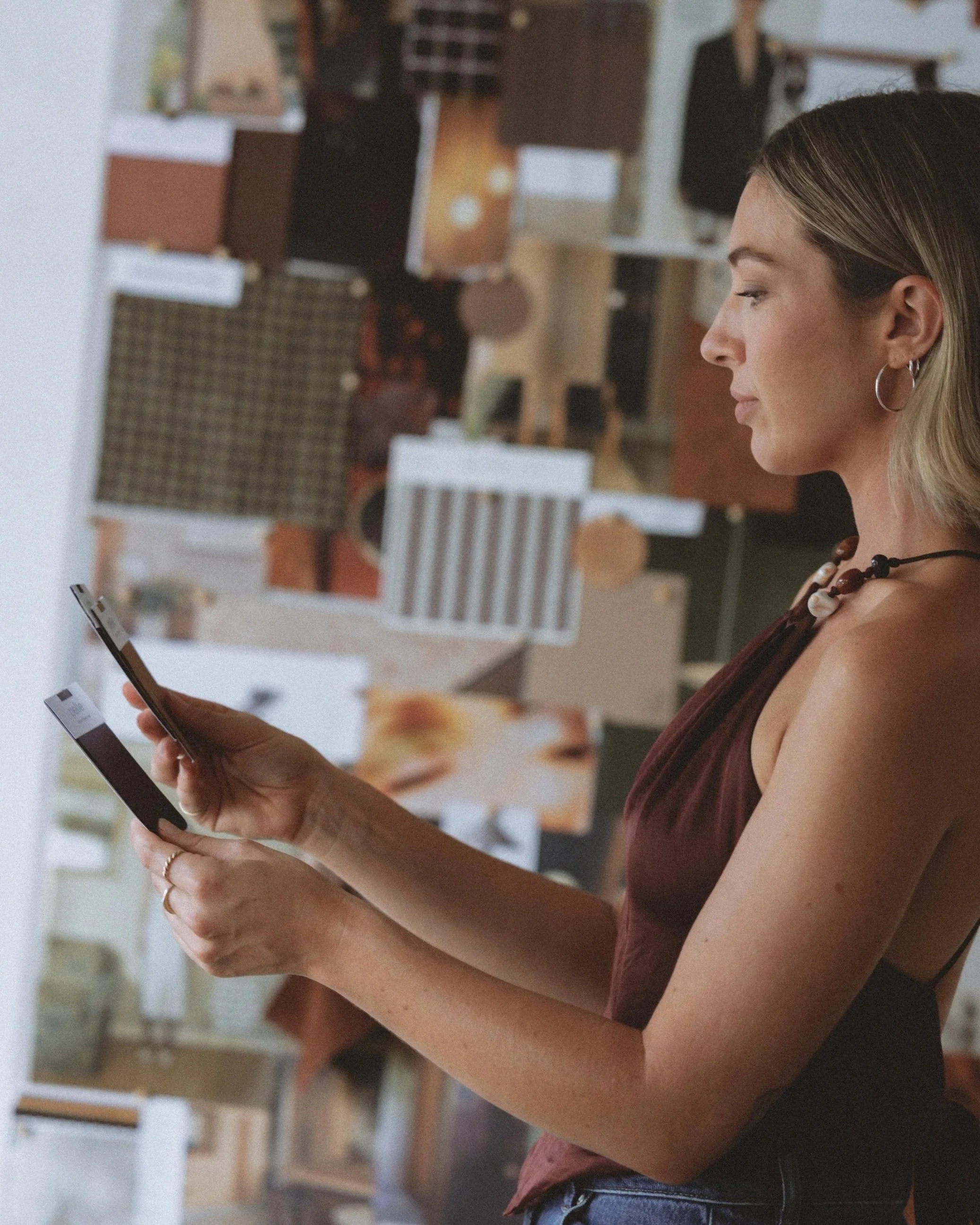 A woman with blonde hair wearing a brown top and multiple necklaces is looking at her phone, sitting at a table in a casual indoor setting.