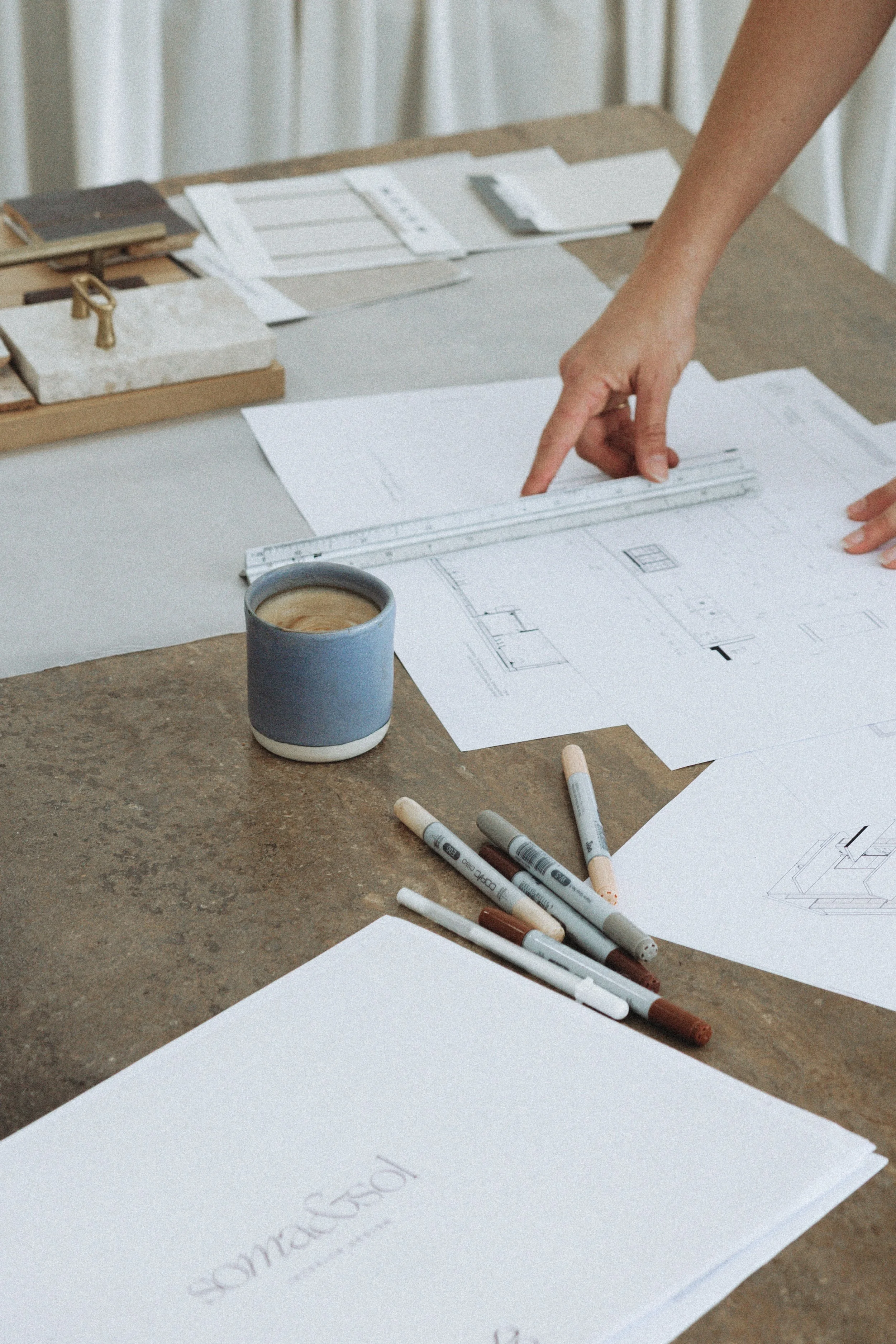 Top view of a workspace with architectural drawings, markers, a ruler, a coffee mug, and a person's hand pointing to a design on paper.