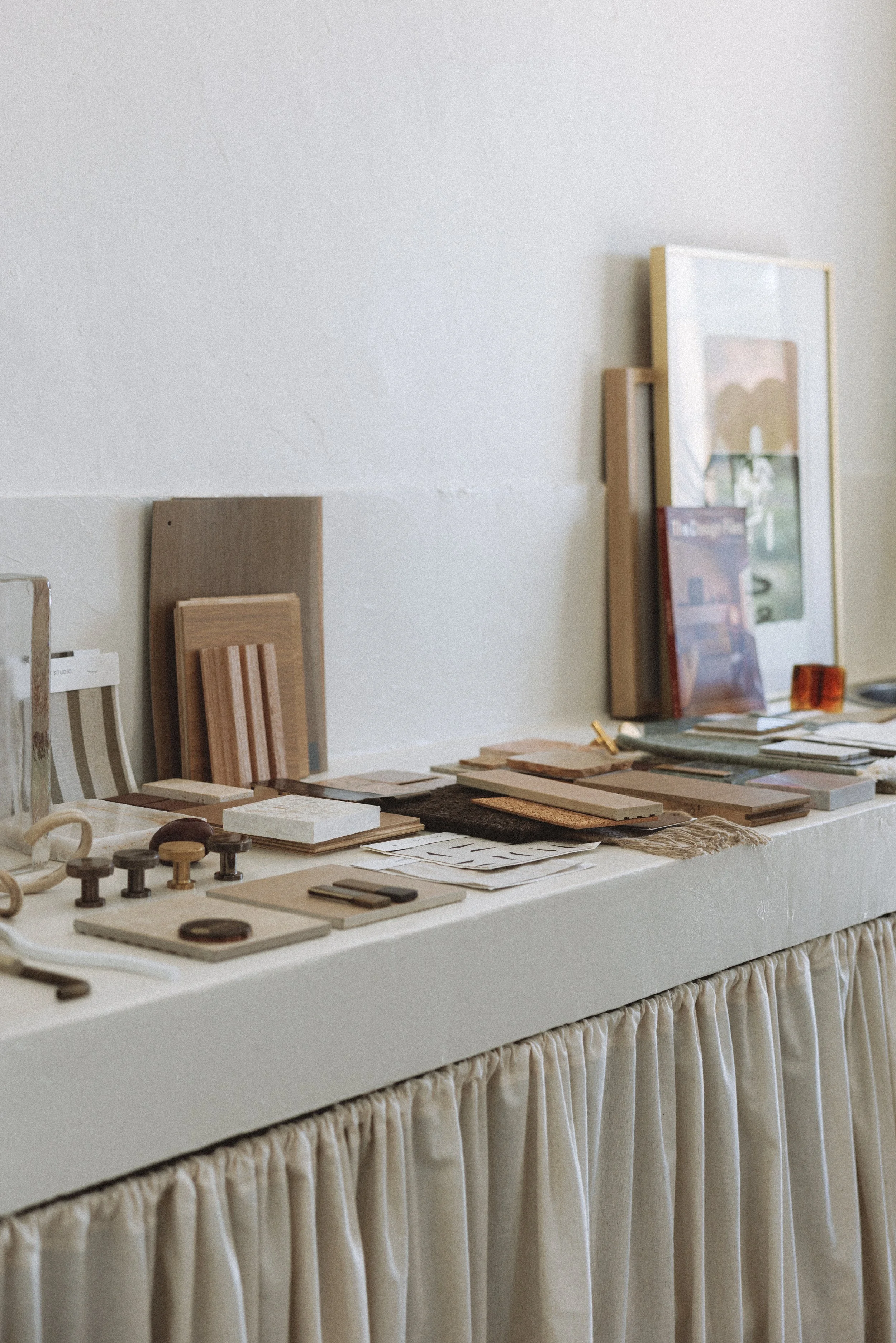 Display of various interior design materials, such as wood samples, tiles, and color swatches, arranged on a table with a white ruffled tablecloth, and framed artworks leaning against the wall in a neutral-colored room.