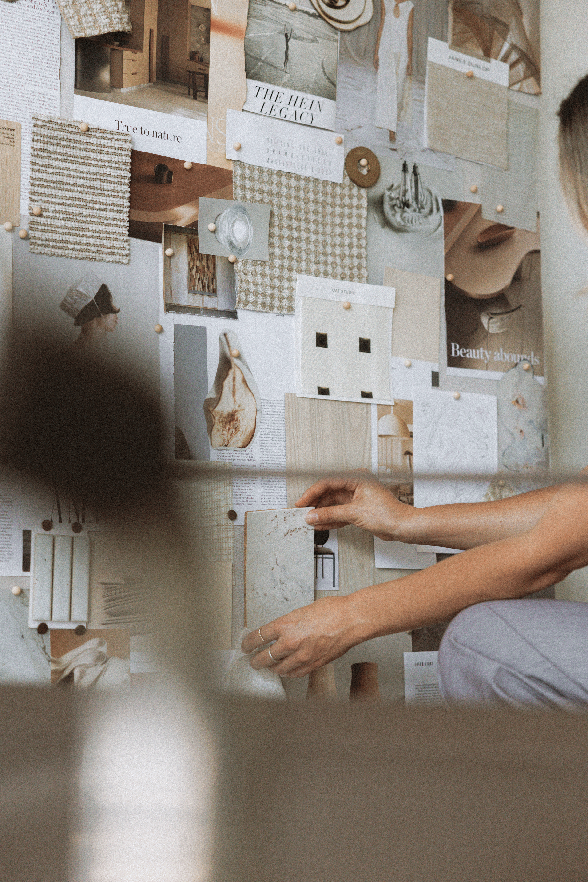 A person sitting and working on a collage or mood board made of various paper, fabric, and magazine clippings pinned to a wall. The collage includes images, text, fabric samples, and sketches in neutral tones.