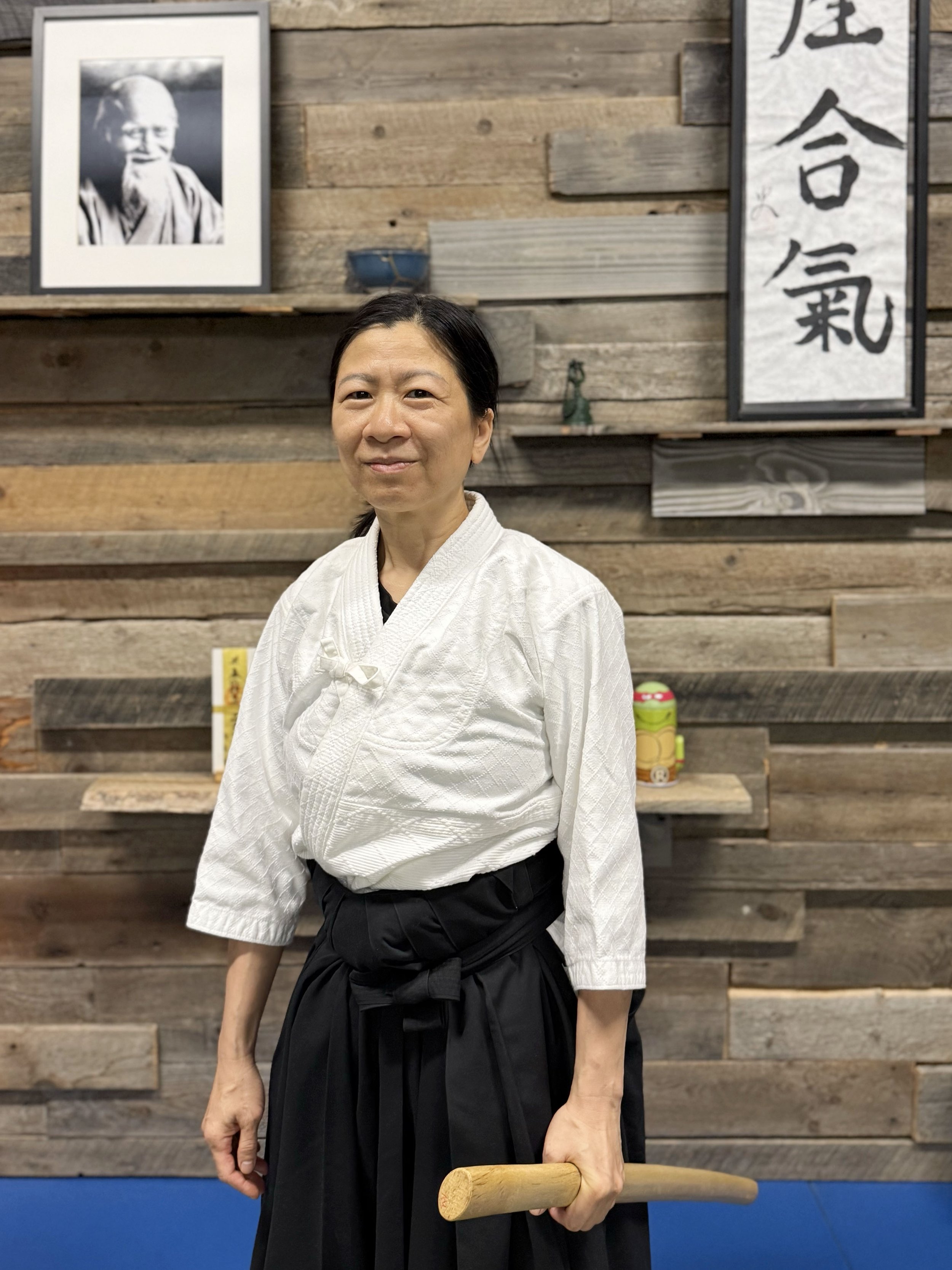 A woman dressed in a martial arts uniform holding a wooden sword, standing in front of a wooden wall with framed Japanese calligraphy and a black and white portrait of an elderly man.