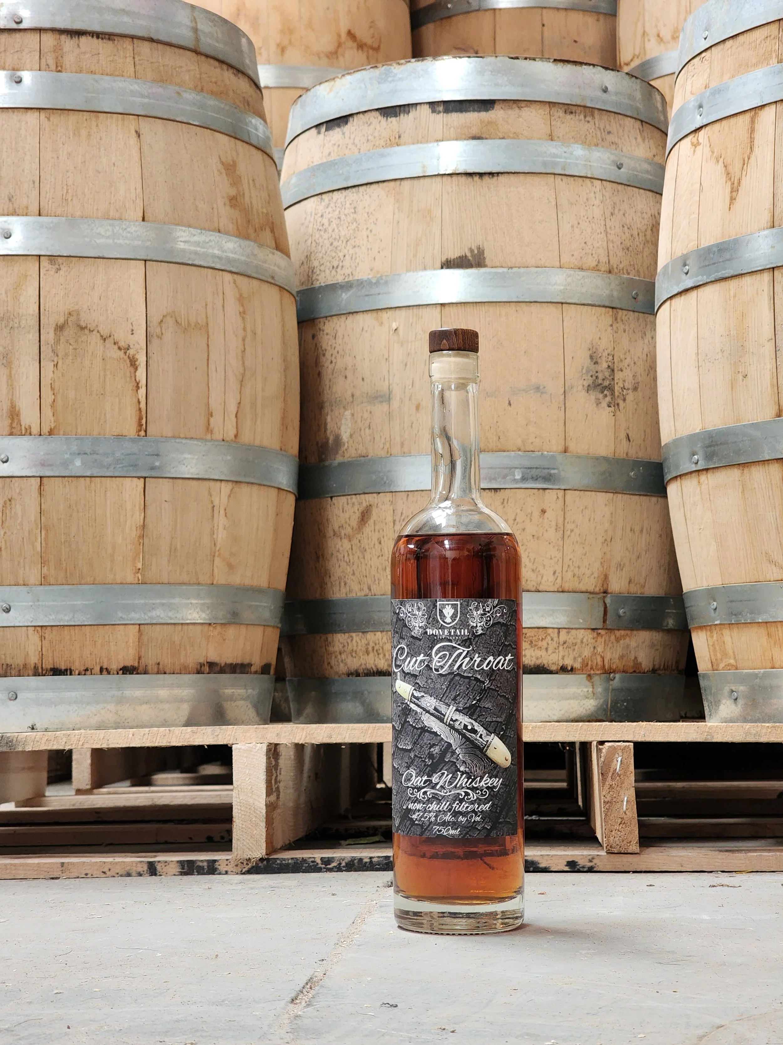 A bottle of whiskey labeled 'Cut Throat' stands on a concrete floor in front of stacked wooden barrels.