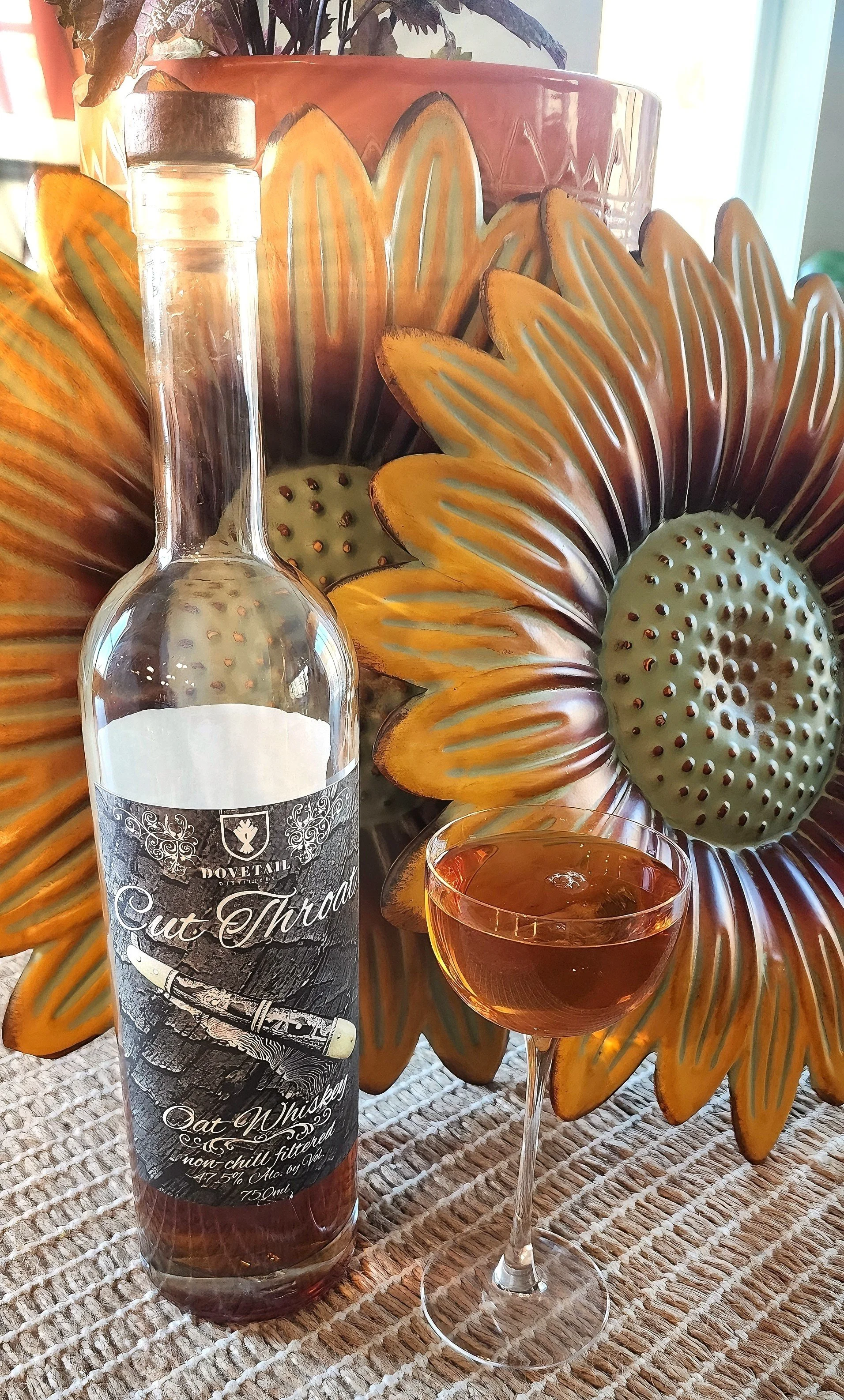 A bottle of Cut Throat oat whiskey with a whiskey cocktail, set on a woven surface in front of large sunflower wall decor.